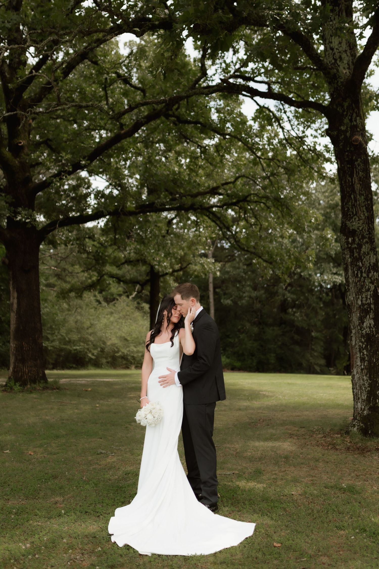 Bride and groom share a romantic kiss beneath tall oak trees in a lush green outdoor setting.
