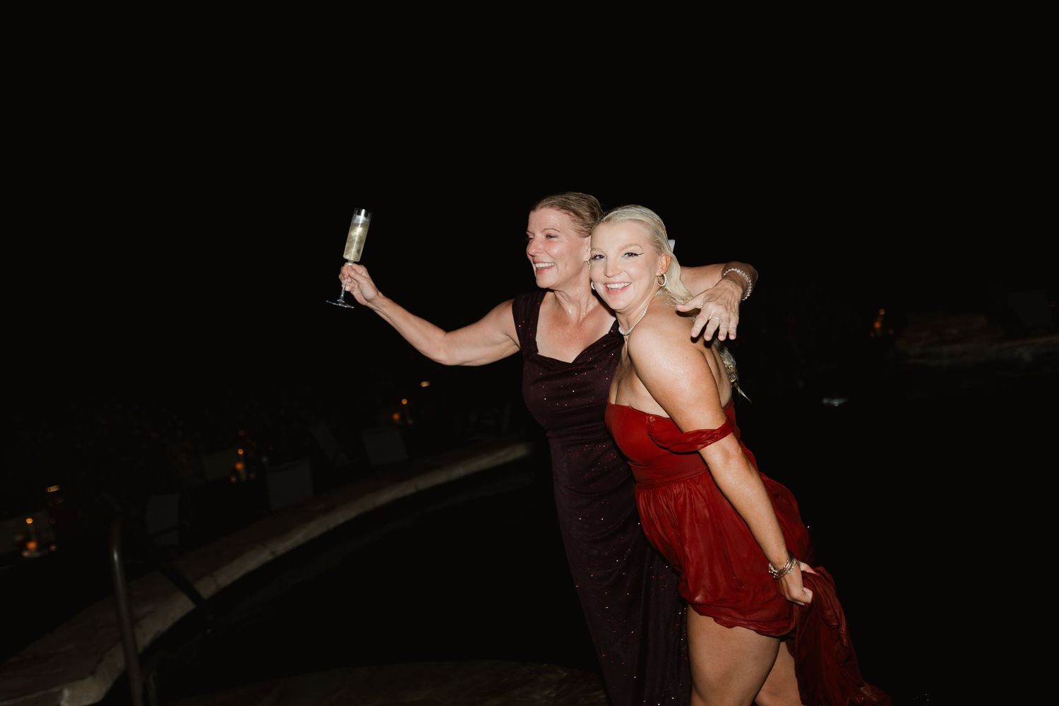 Two women in elegant dresses celebrate at a night event, one holding a champagne glass for a selfie.