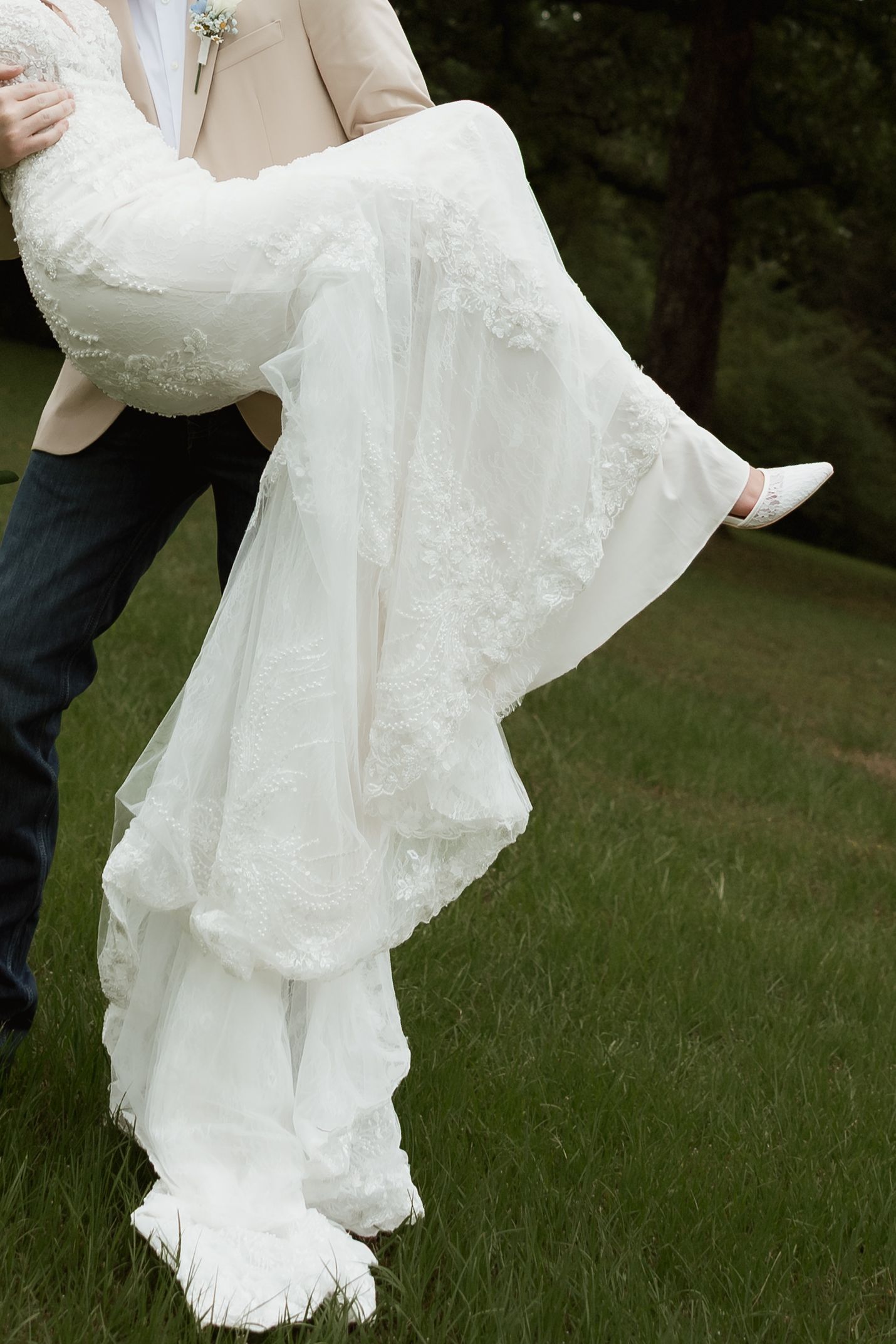 Groom in tan blazer carries bride in white lace wedding dress on green lawn.