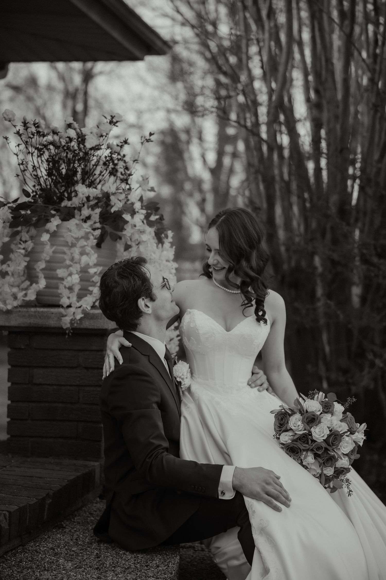 Black and white romantic wedding photo of groom kneeling before bride holding bouquet near floral arch.