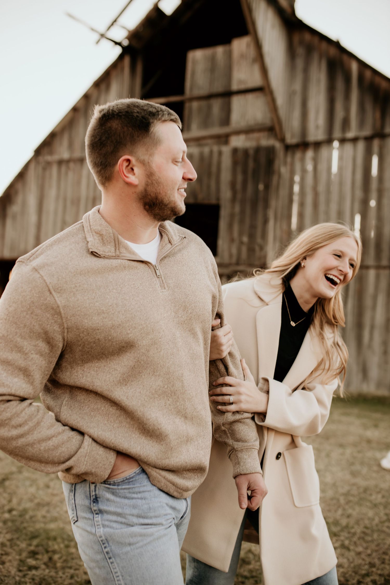 Laughing couple in neutral toned outfits holding hands near rustic wooden barn during outdoor photo session.