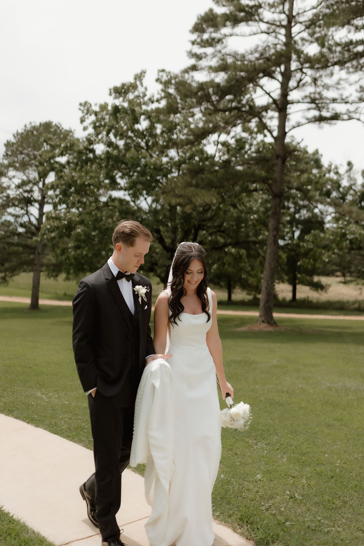 Bride and groom walking on pathway, groom in black tuxedo and bride in white gown holding bouquet.