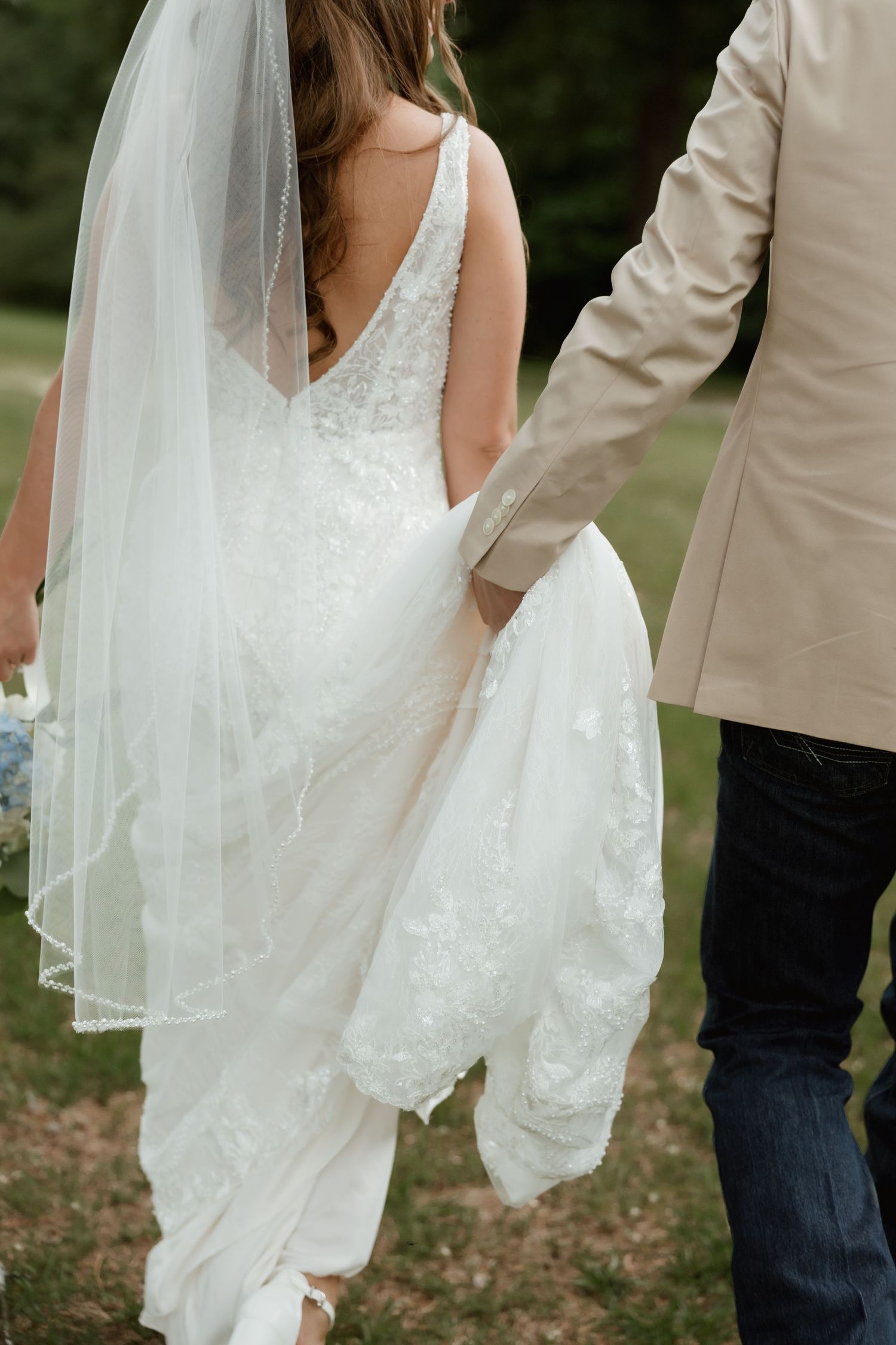 Bride in lace wedding gown and veil walks hand in hand with groom outdoors.