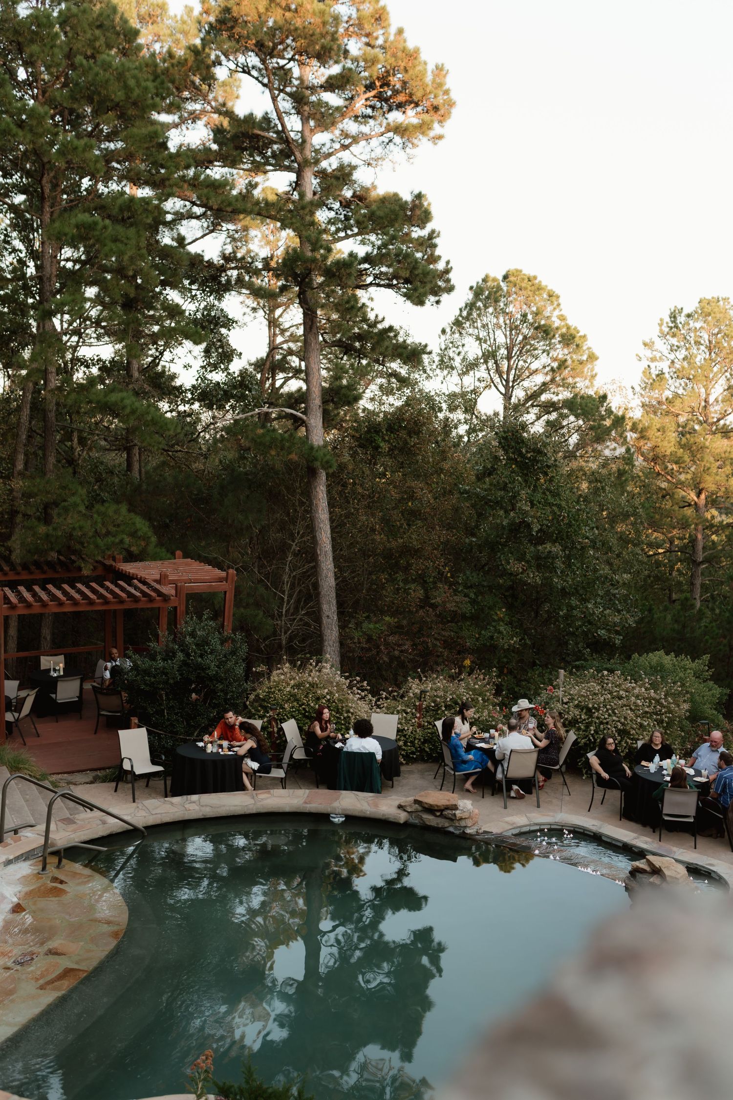 Wedding guests dining poolside at an elegant outdoor reception surrounded by tall pine trees.