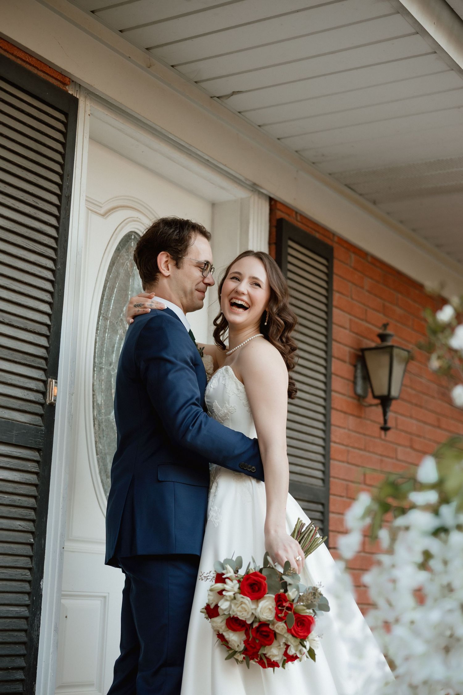Smiling bride holding red and white bouquet embraces groom in navy suit on home porch.