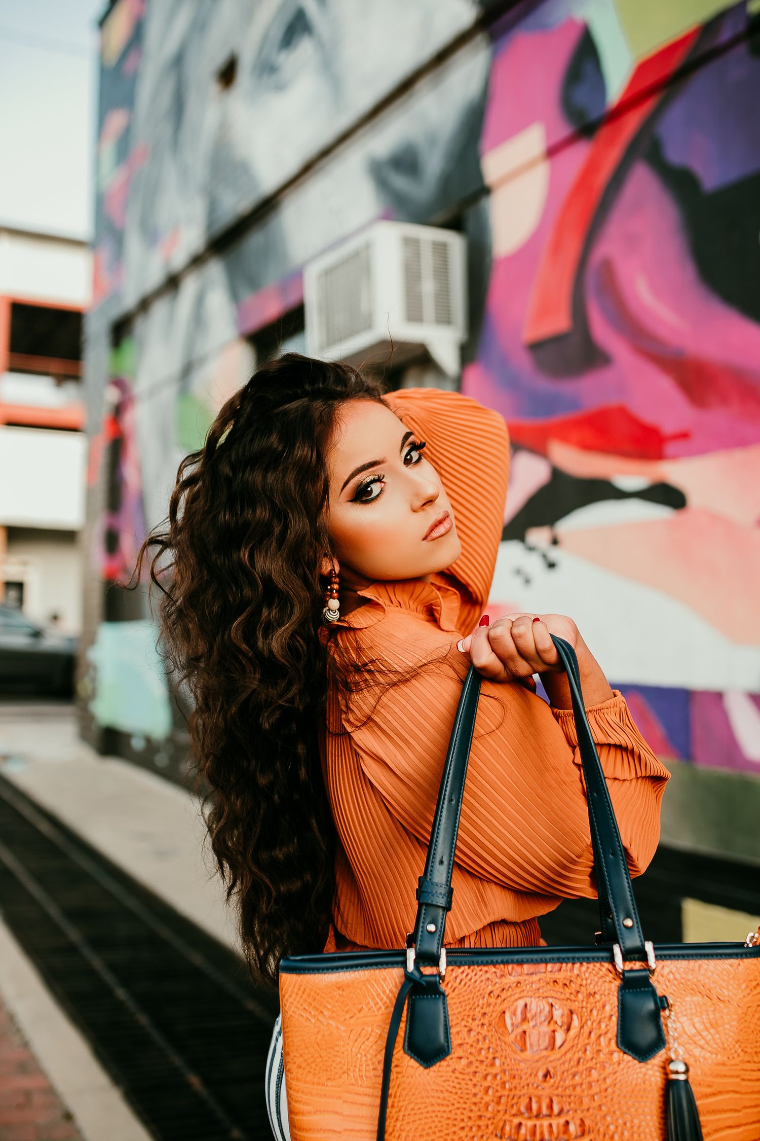 Woman in orange outfit holding matching handbag posing before colorful graffiti wall.