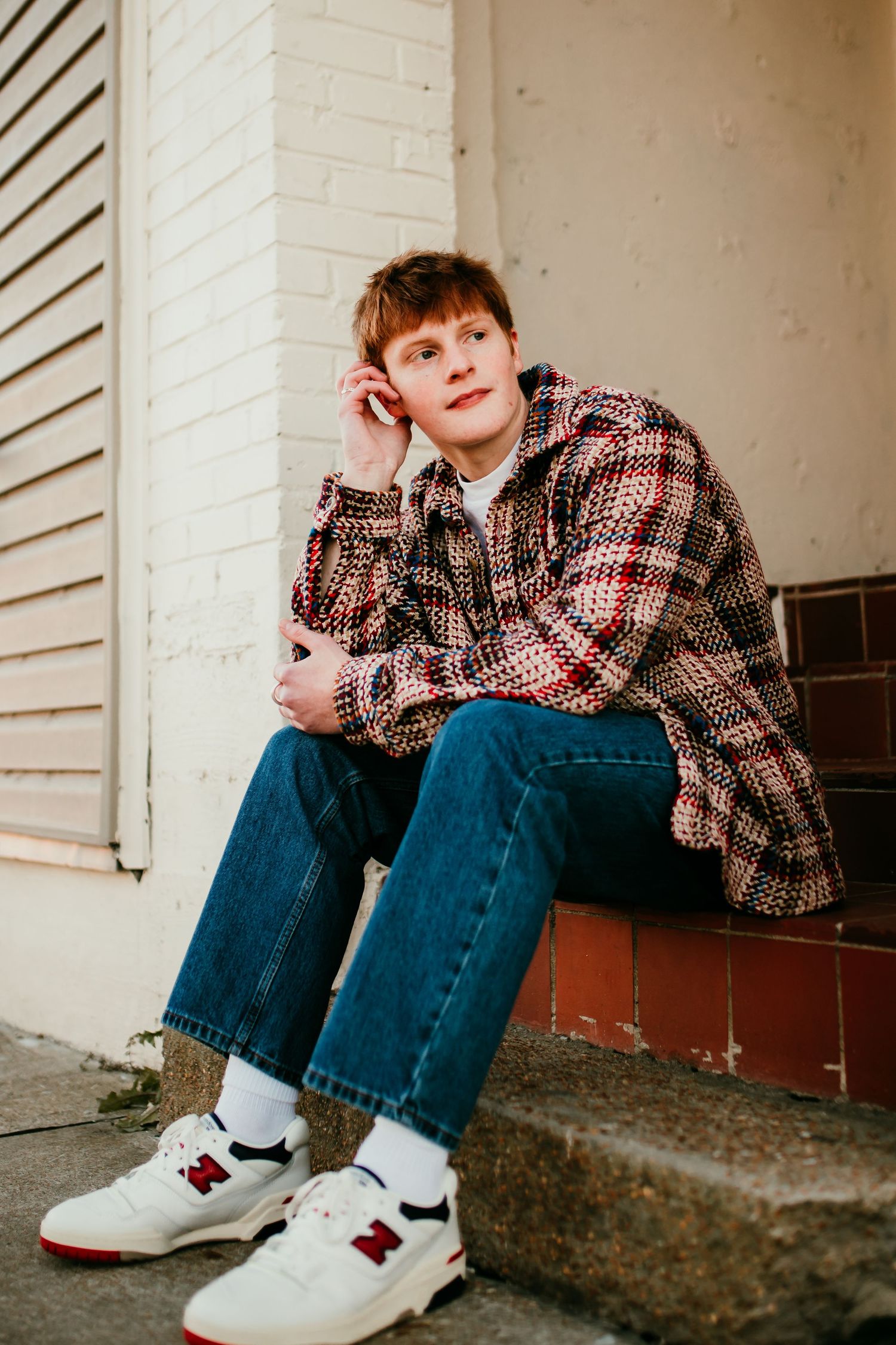 Young man in plaid shirt and jeans sitting on steps wearing New Balance sneakers.