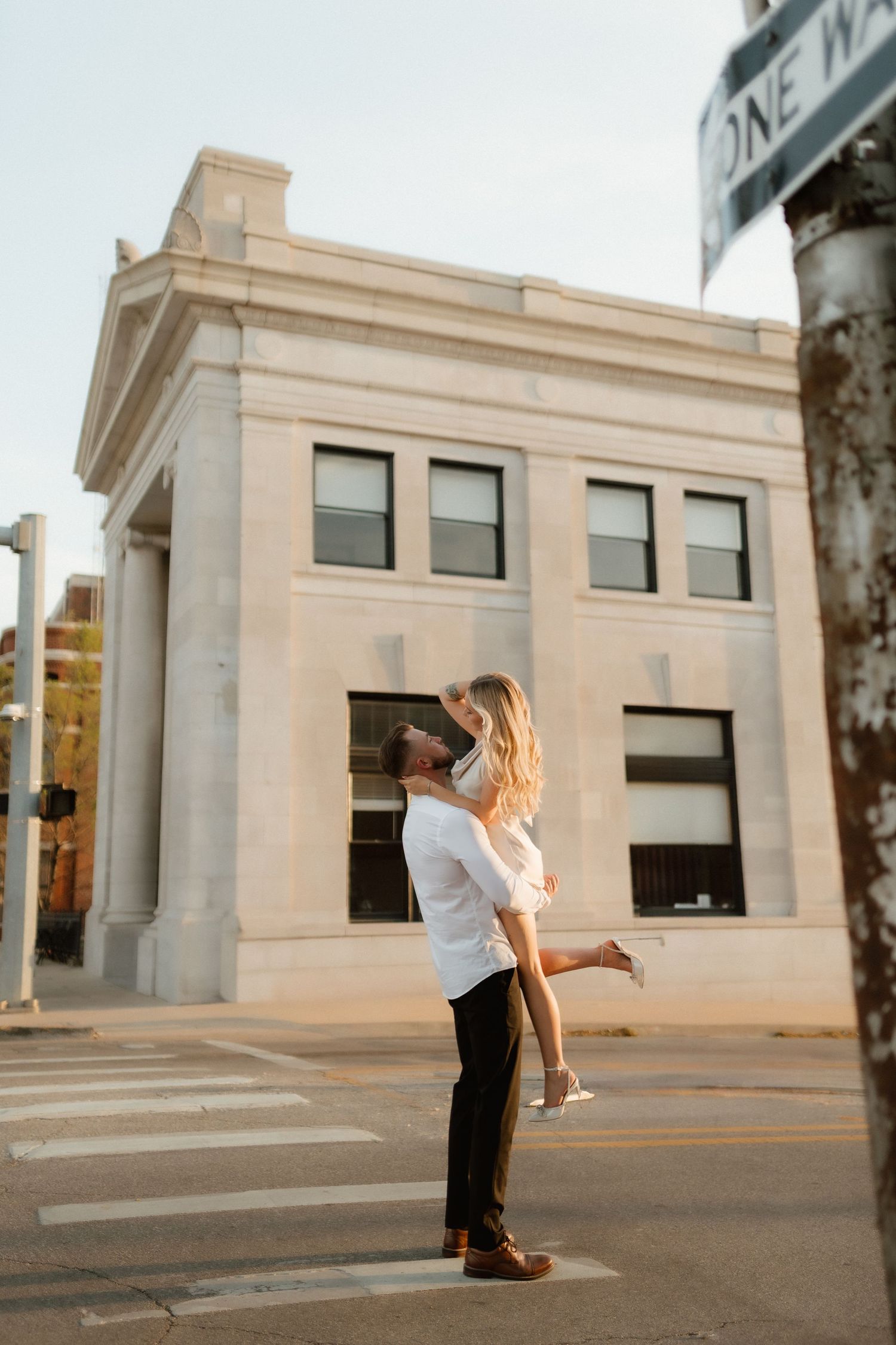 A couple shares a romantic lift embrace at a city crosswalk near a One Way sign.