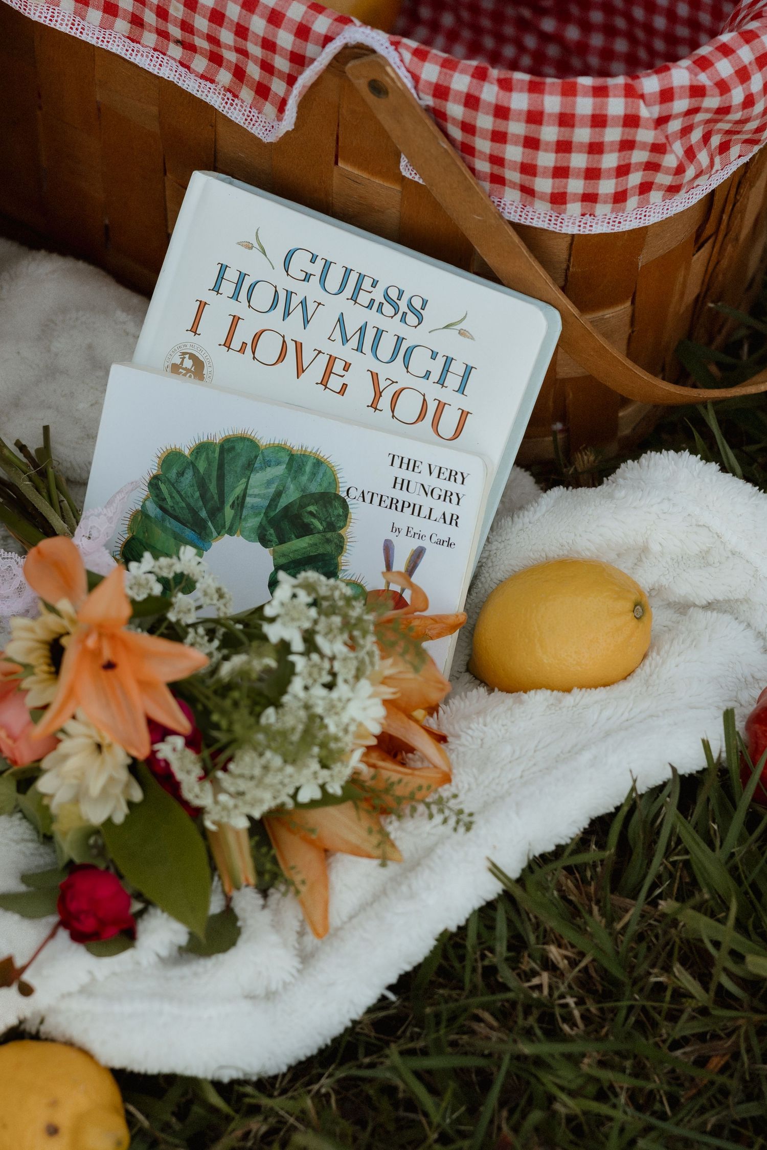 Children's books, wildflowers, and lemons arranged on white blanket beside wicker picnic basket.
