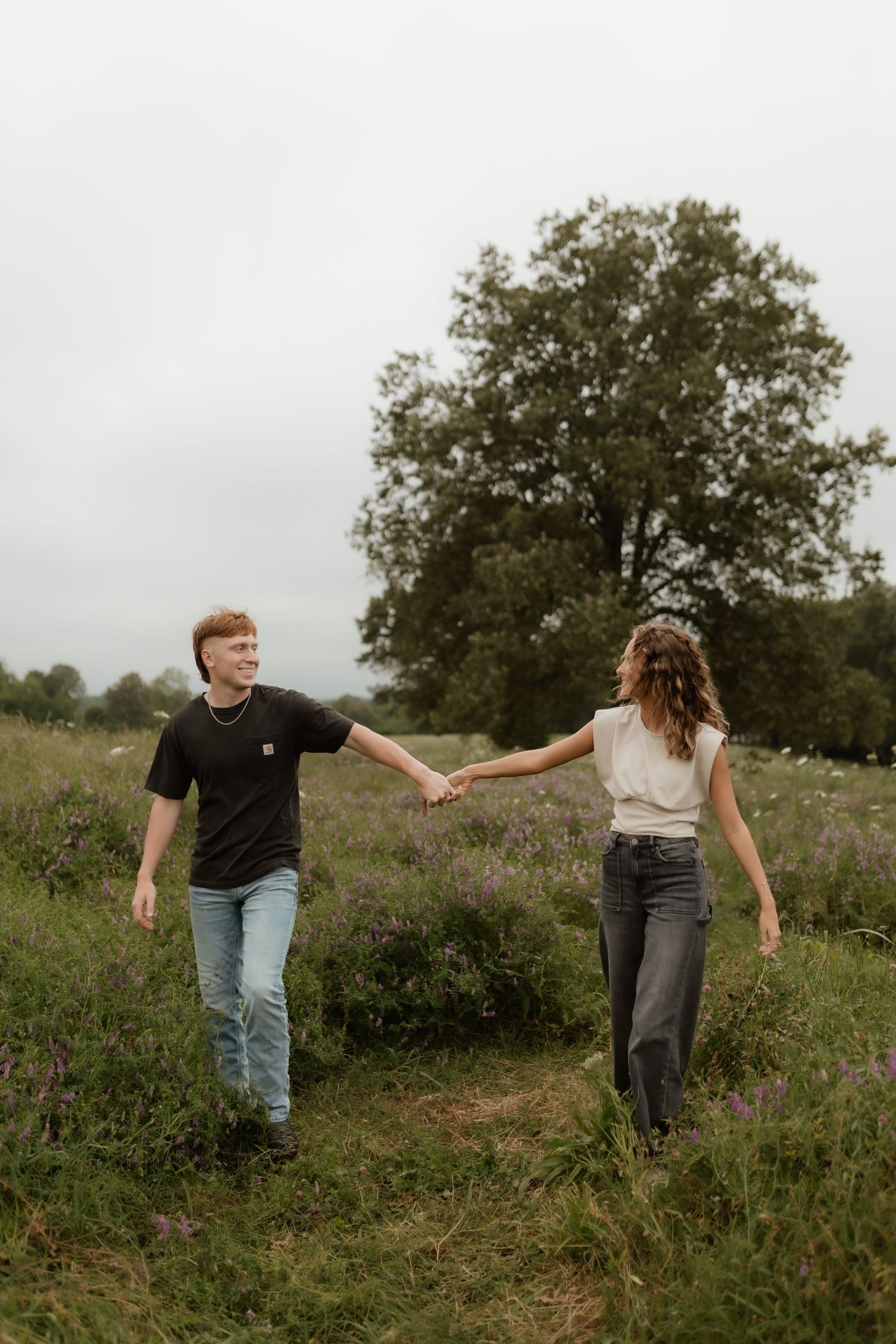 Young couple holding hands while walking through a lush green meadow with wildflowers and a large tree.