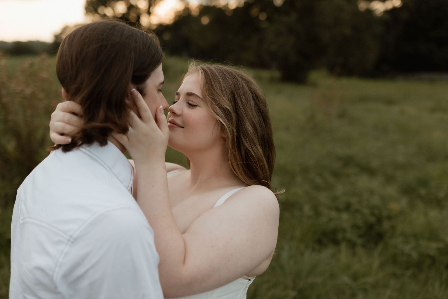Couple sharing a romantic kiss in golden hour sunlight in a lush green field, woman in white dress.