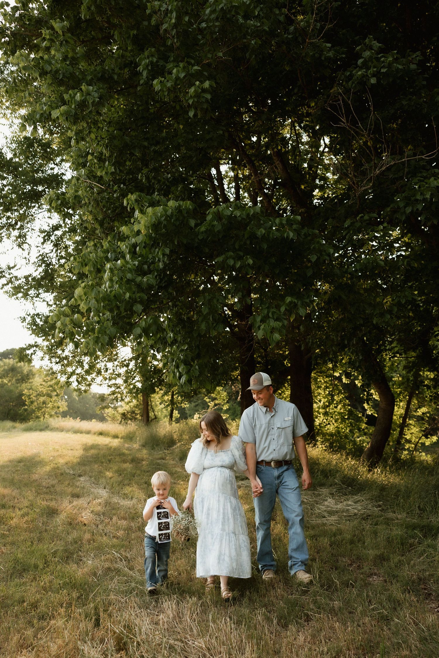 Family of three walking in sunlit meadow, child holding ultrasound photo near tall green trees.