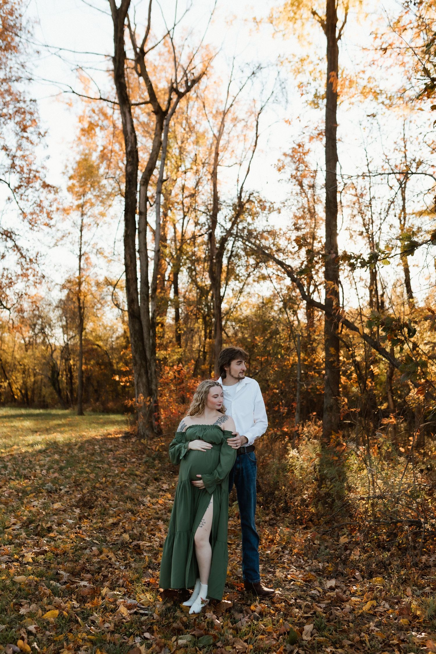 Pregnant woman in green maternity gown embraced by partner in autumn forest during golden hour.