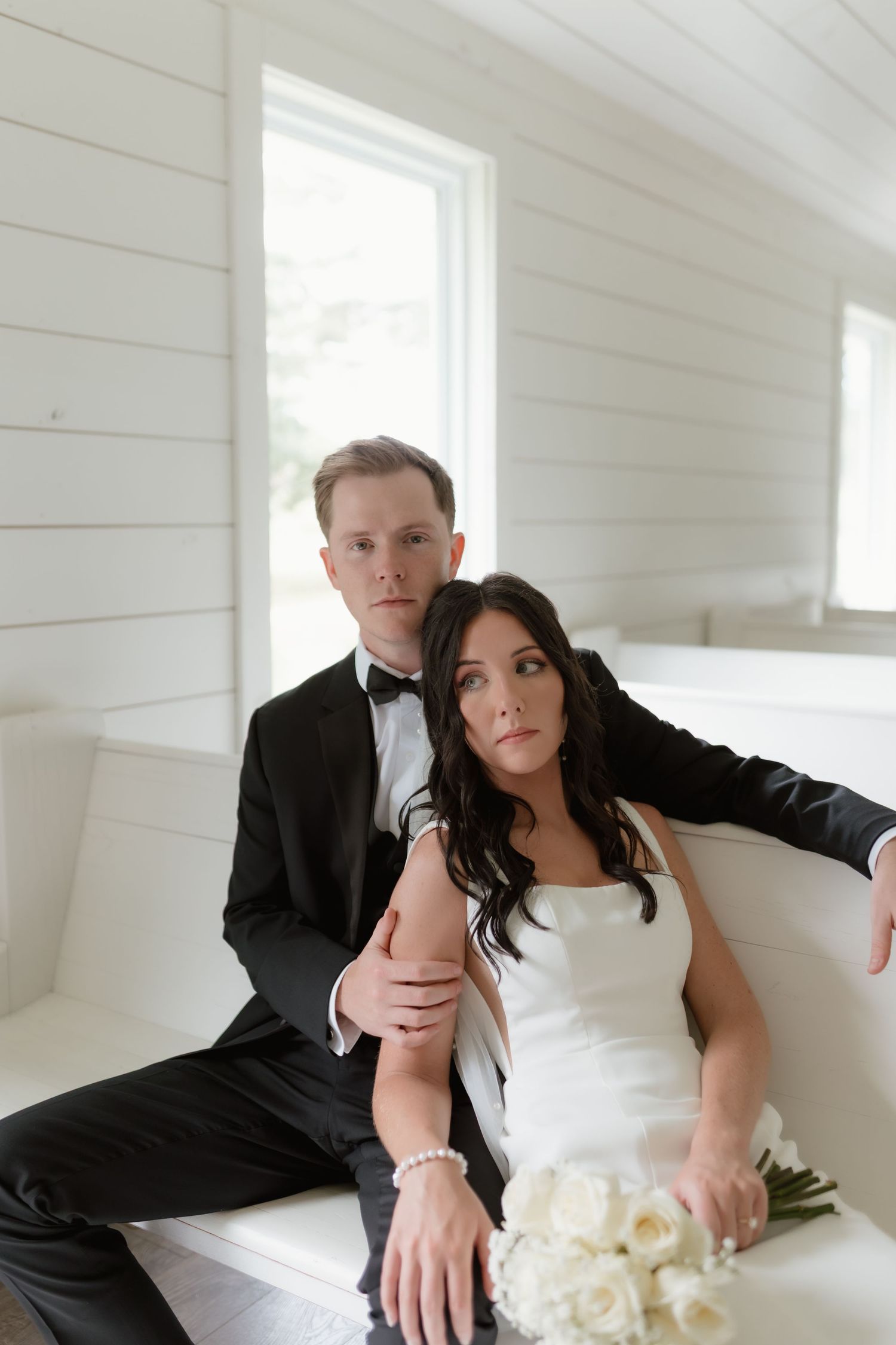 Elegant bride and groom posing on white couch indoors, groom in black tuxedo, bride holding white roses.