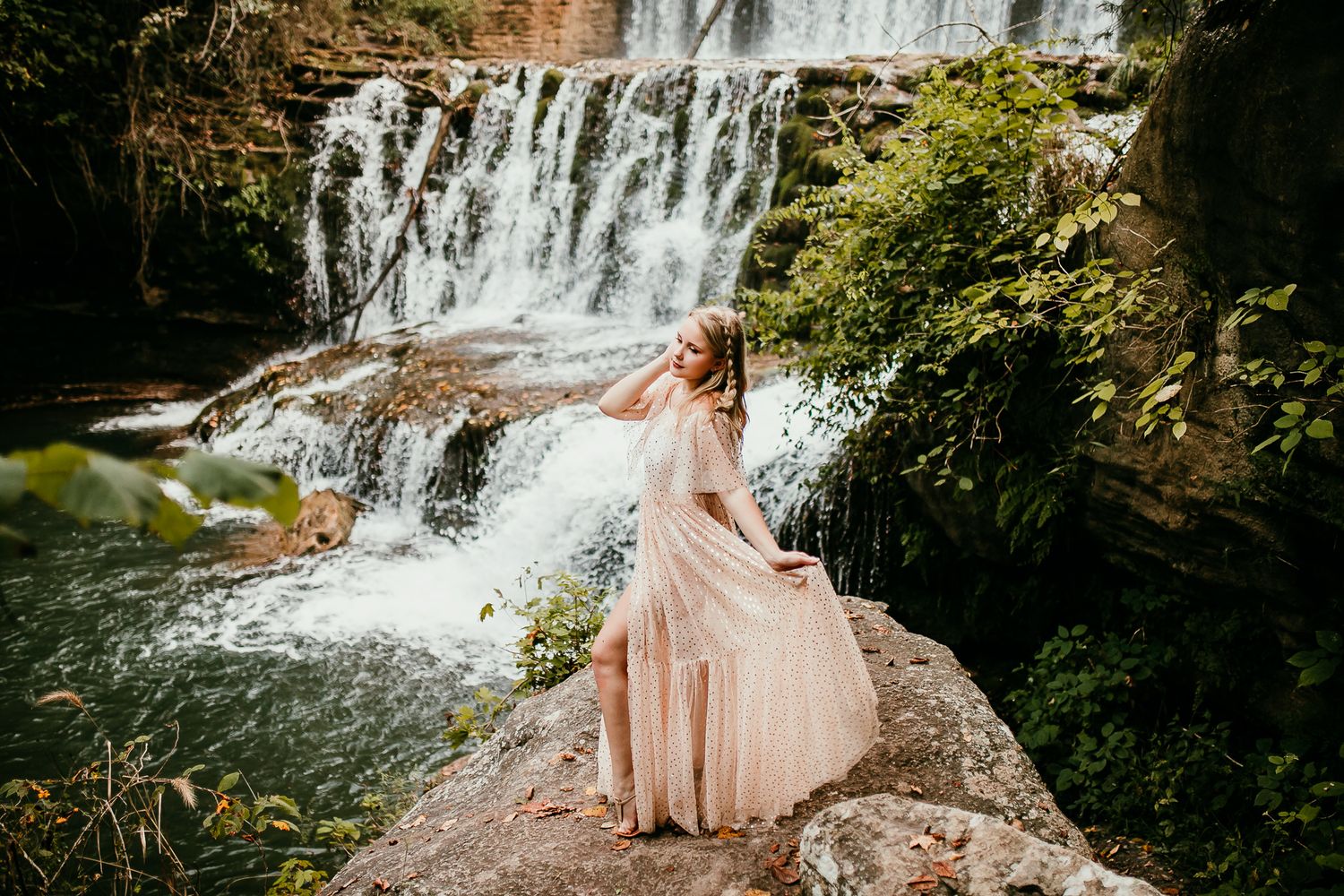 Blonde woman in flowing blush gown twirls on rock with dramatic waterfall backdrop in nature.