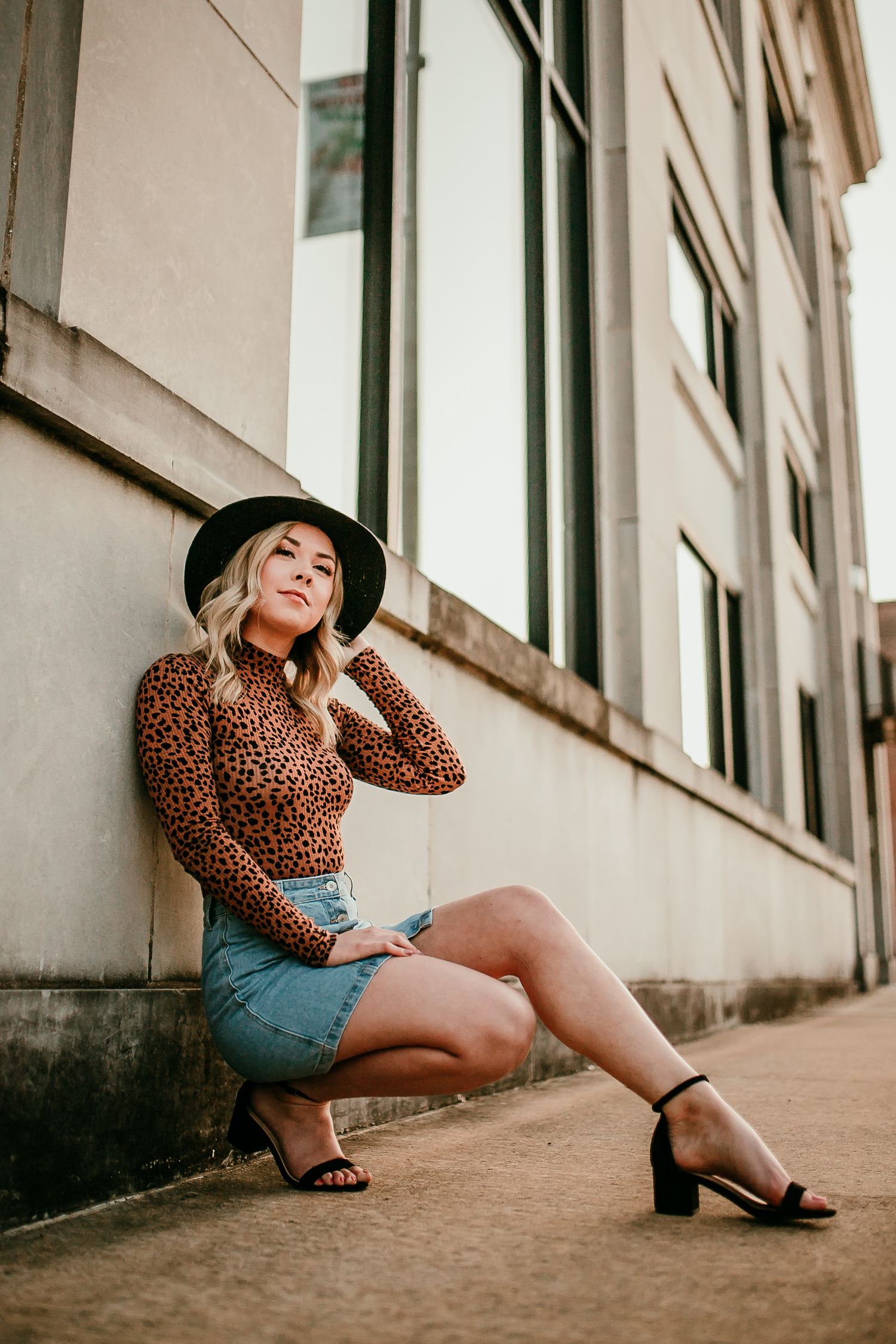 Young woman in leopard print top, denim skirt, and black wide-brim hat poses against urban building wall.