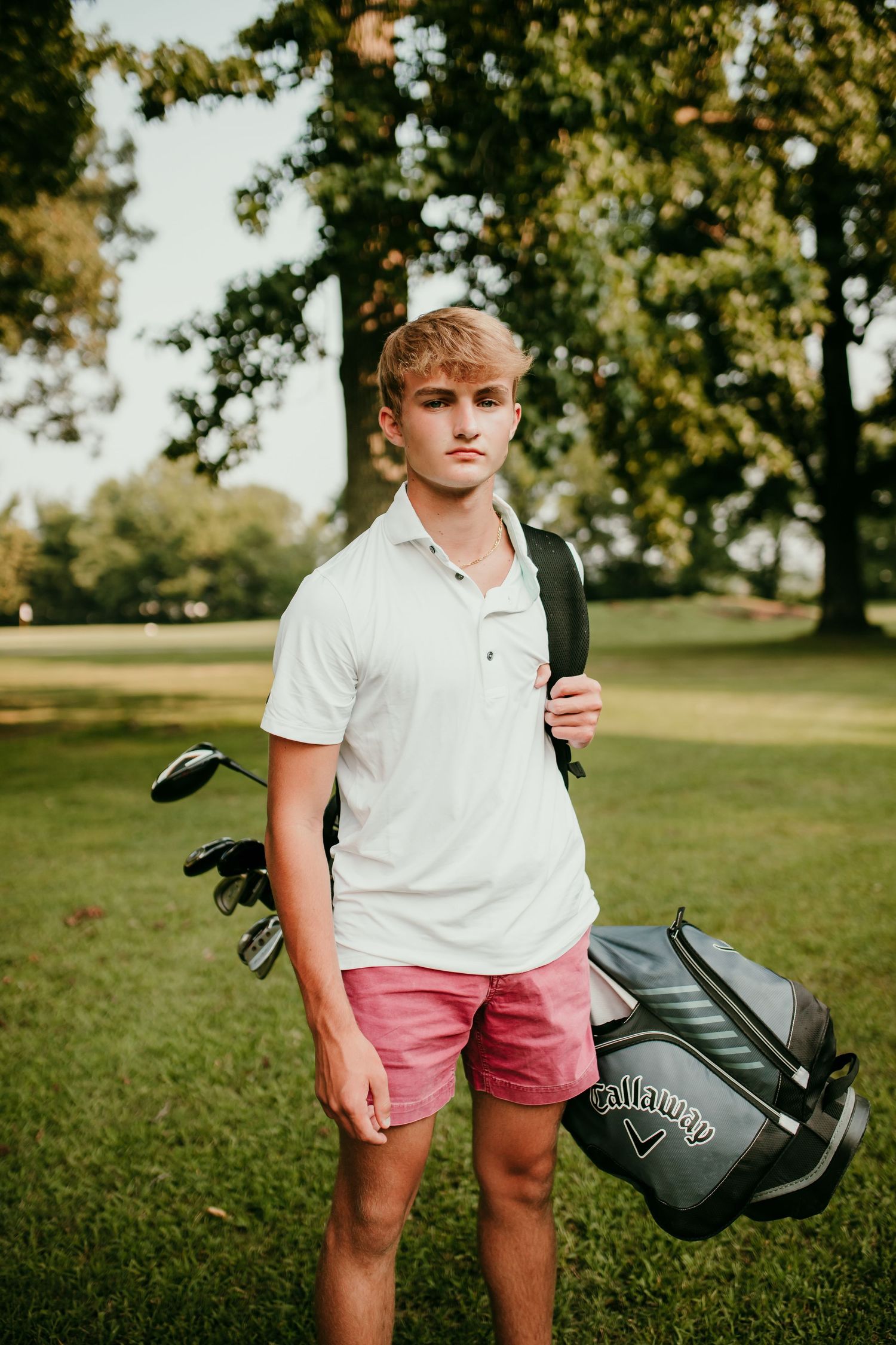Teen boy in white polo and pink shorts carries Callaway golf bag on green golf course.