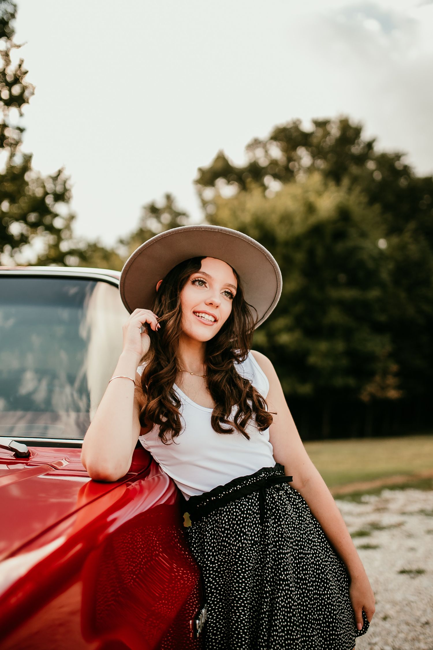 Brunette woman in white tank, polka dot skirt, and wide-brim hat leans against vintage red truck outdoors.