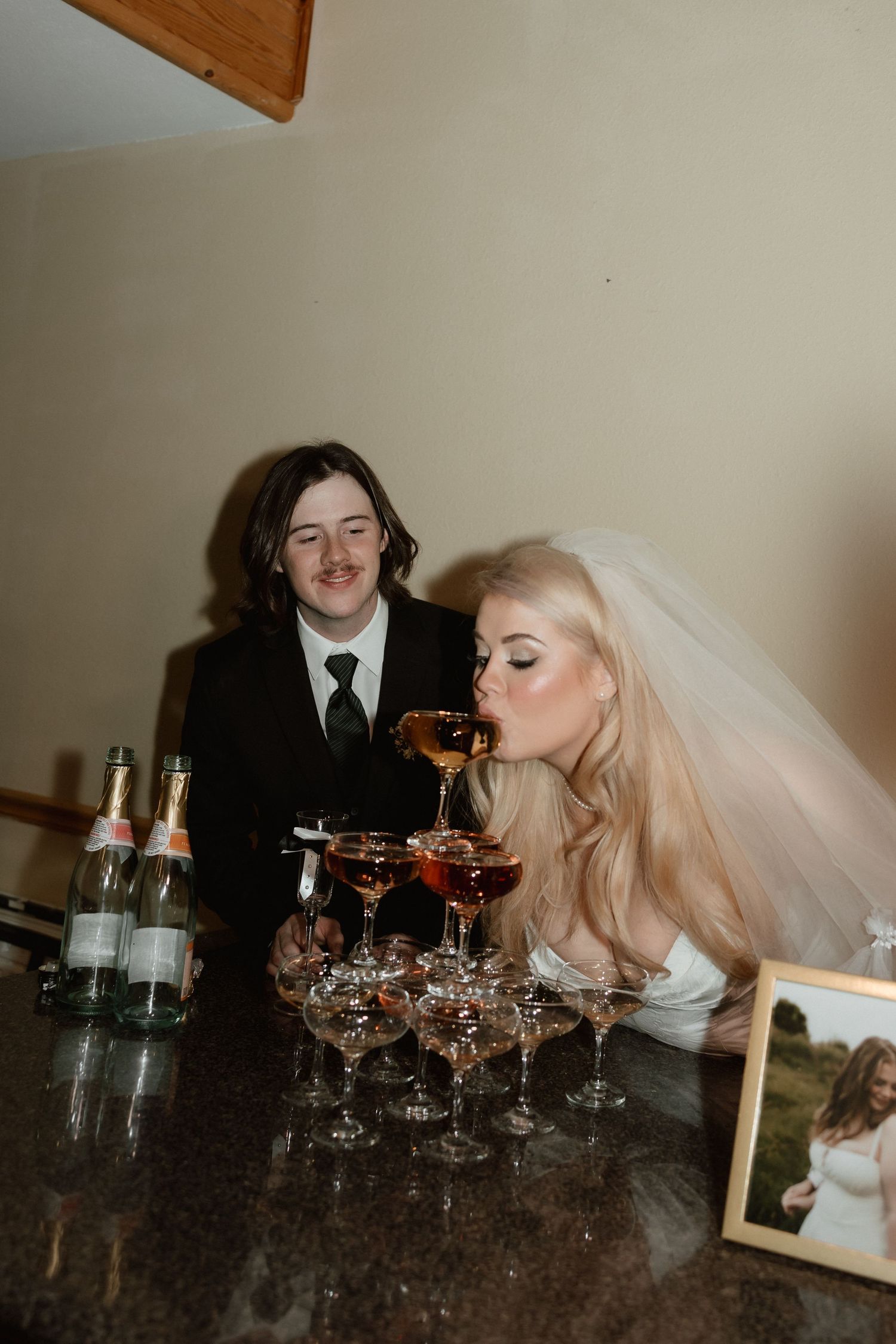 Blonde bride in veil sipping from champagne tower while groom watches at reception.