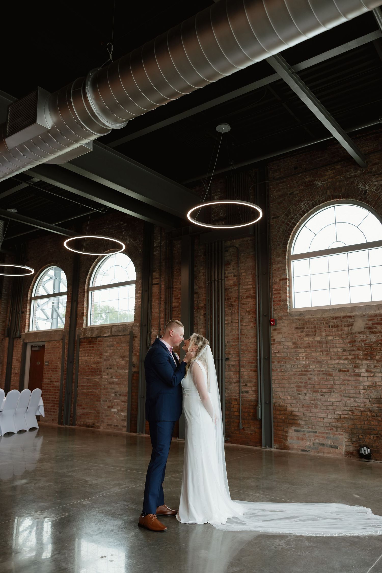 Bride and groom share first kiss in industrial brick wedding venue with circular pendant lights overhead.