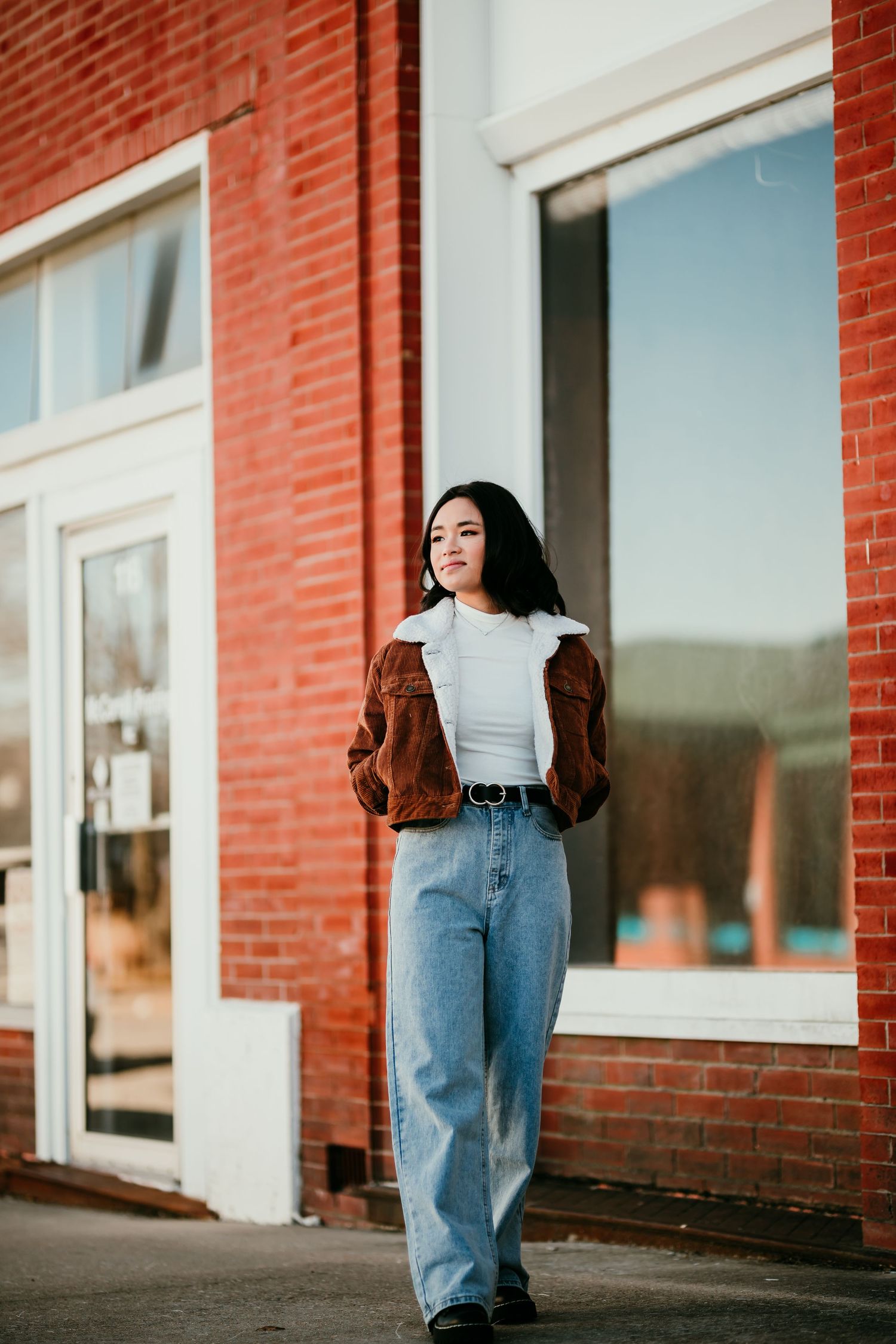 Stylish young Asian woman in brown jacket and wide-leg jeans standing outside a red brick building.