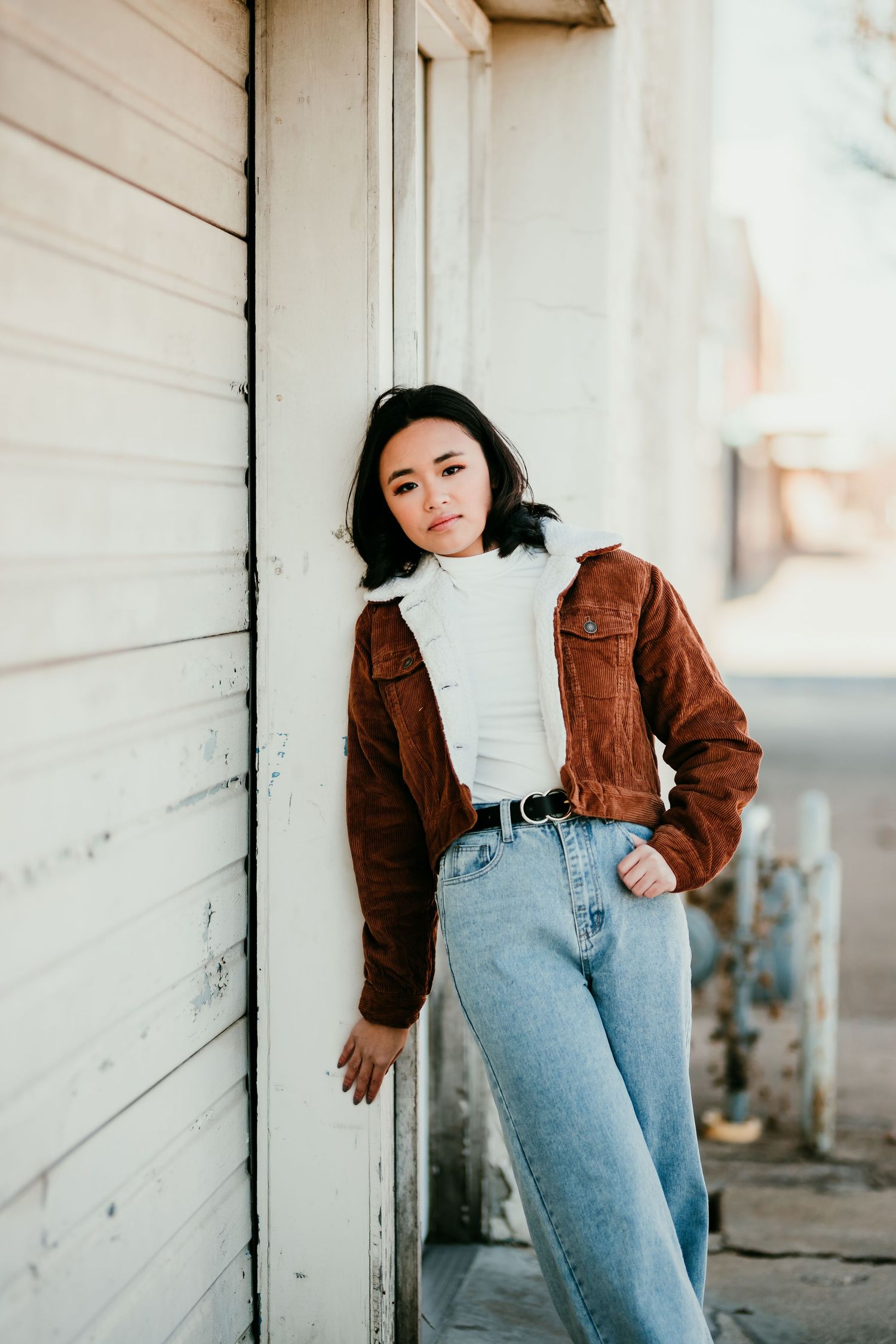 Young Asian woman in brown corduroy jacket and high-waist jeans leaning against white wooden wall outdoors.
