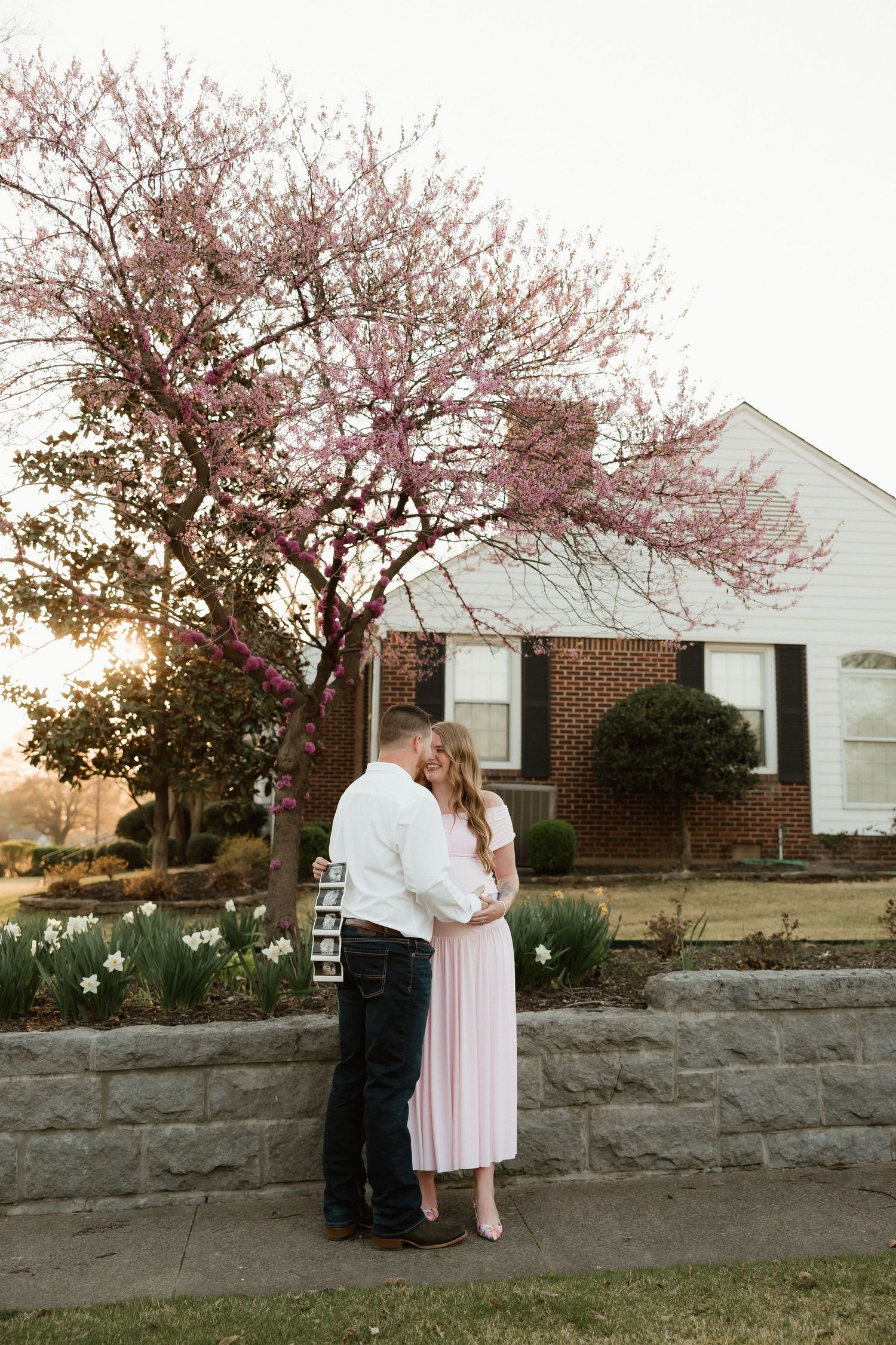 Couple embracing near a blooming pink redbud tree at sunset, man holding ultrasound photos in a maternity session.