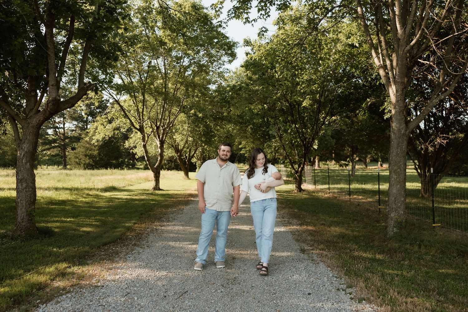 Young couple walking down tree-lined gravel path holding newborn baby in sunny outdoor setting.