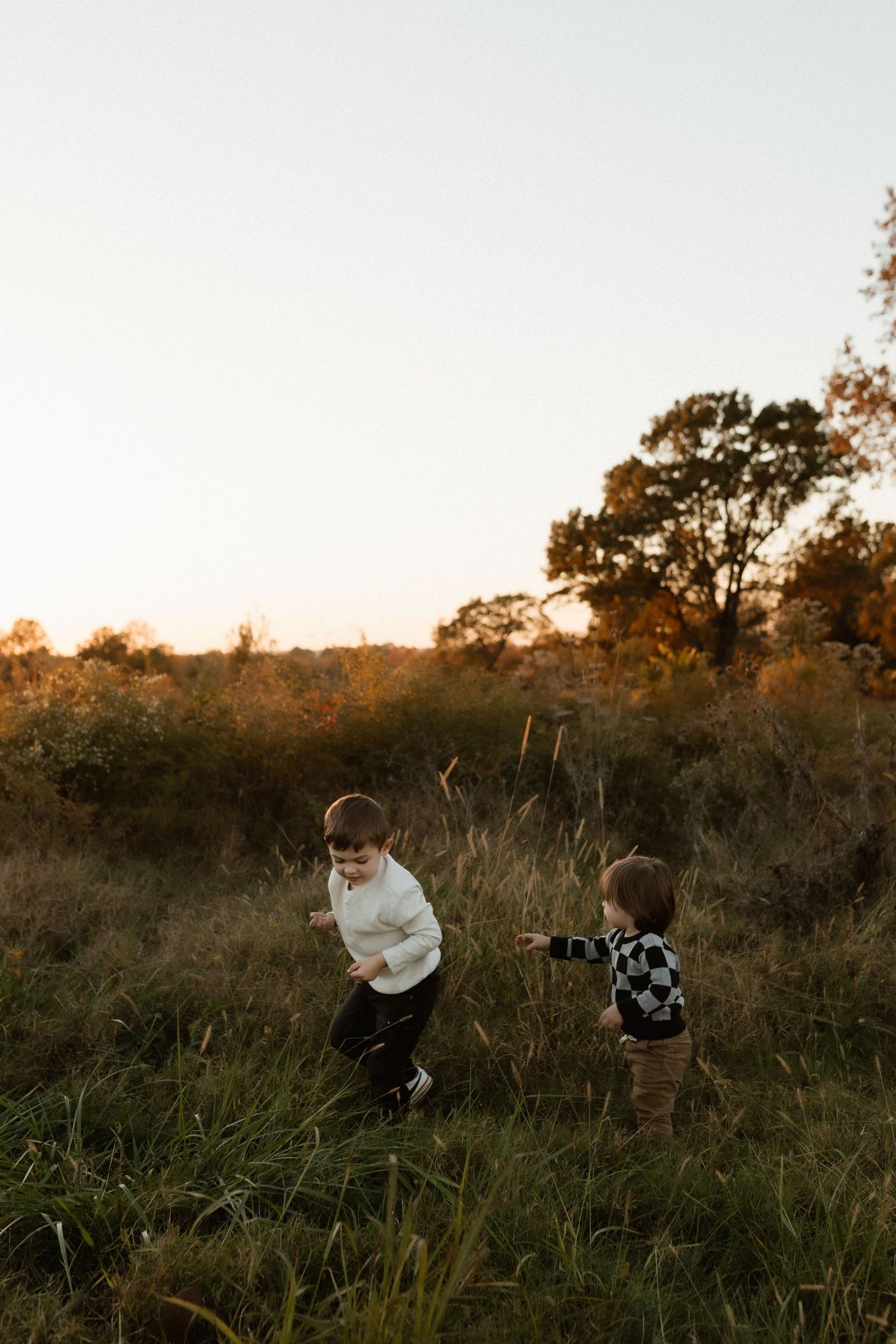 Two young boys in casual outfits play together in tall grass field at sunset.