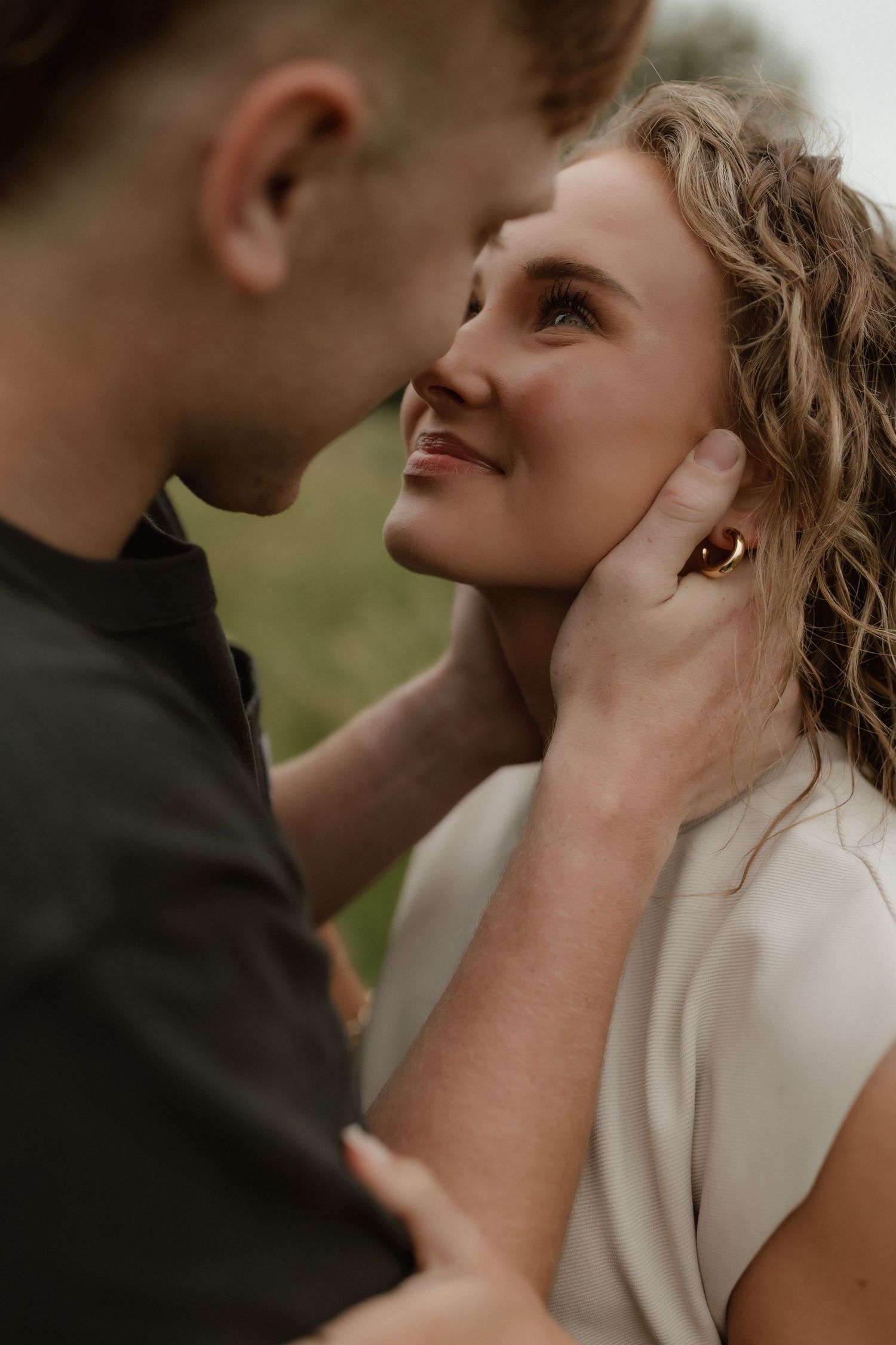 Young woman smiling up at man who gently cups her face during romantic outdoor couples session in green field.