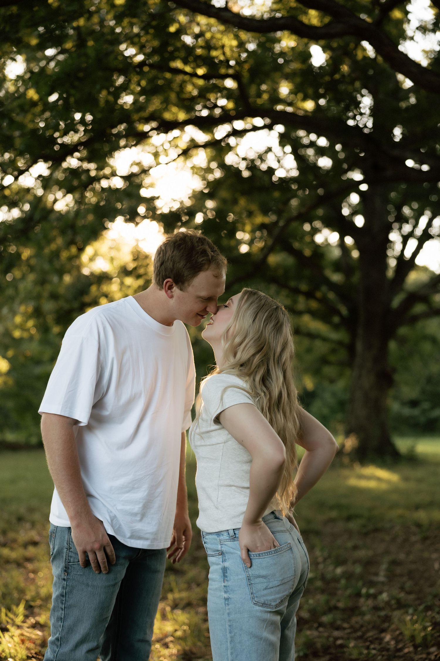 A young couple sharing a tender kiss outdoors in a sun-dappled park with large trees in the background.