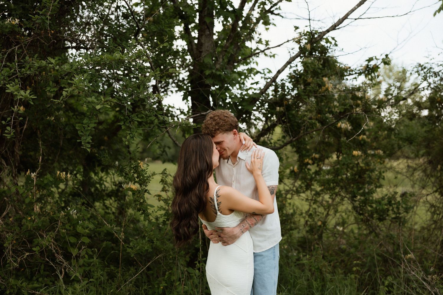 A couple shares a tender embrace near lush green trees in an outdoor natural setting.