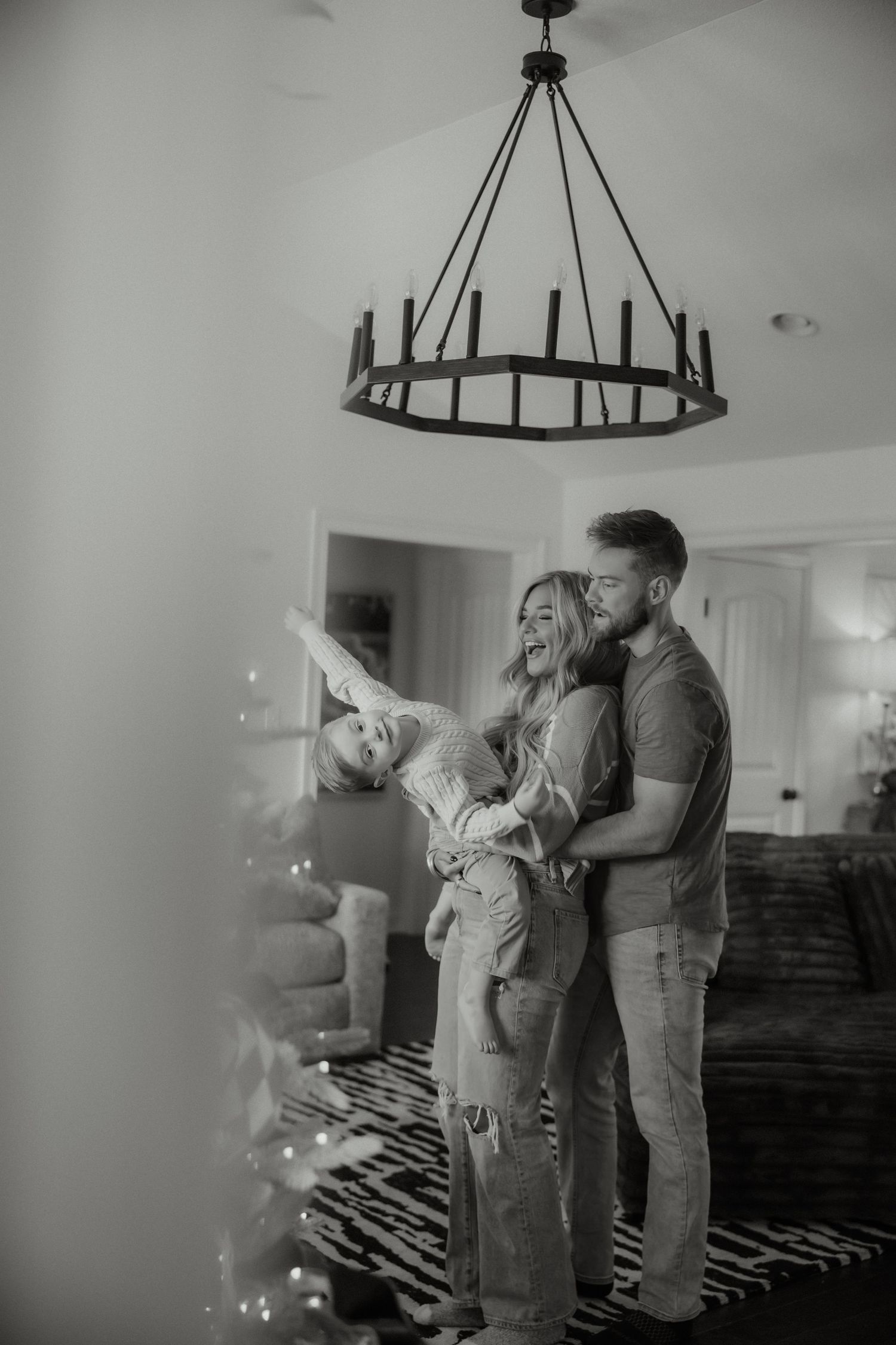 Black and white photo of joyful family with toddler laughing under rustic chandelier near Christmas tree.
