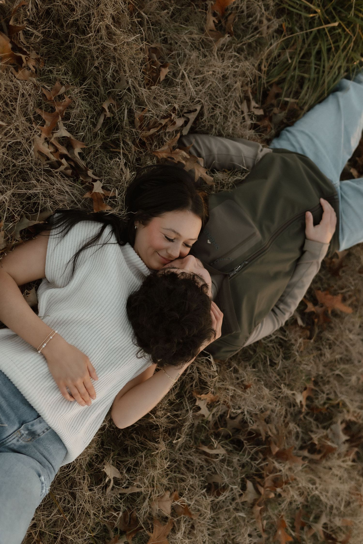 A couple lies together on dry grass and fallen leaves in a cozy autumn embrace.