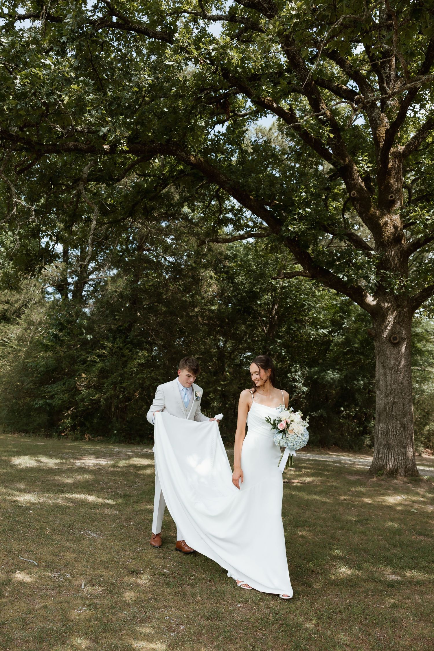 Groom carrying bride's white gown train as couple walks through lush green outdoor setting.