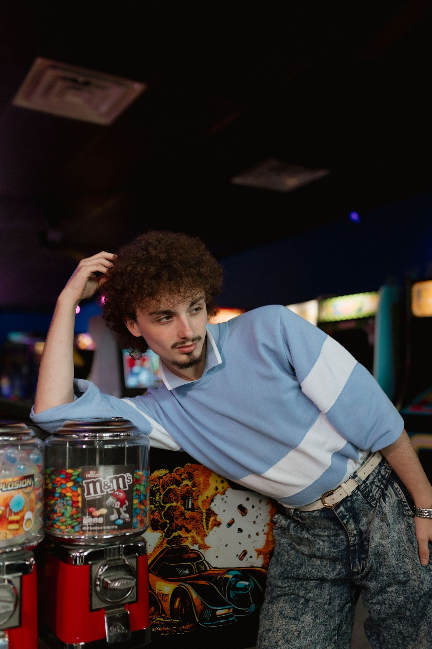Curly-haired man in retro blue sweatshirt leaning on M&M gumball machine in arcade.