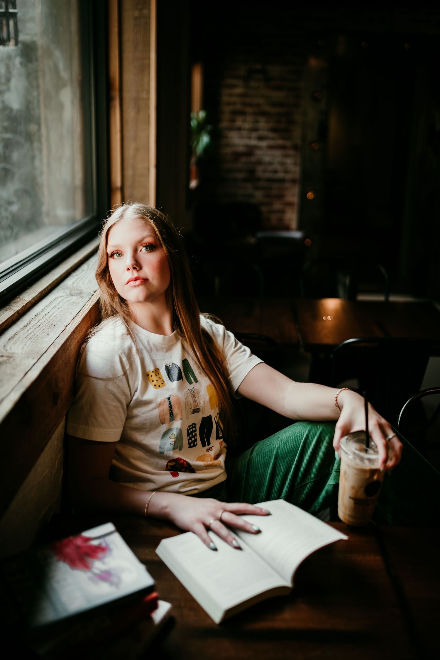 Young woman with long blonde hair sits at cafe table with open book and iced coffee near window.