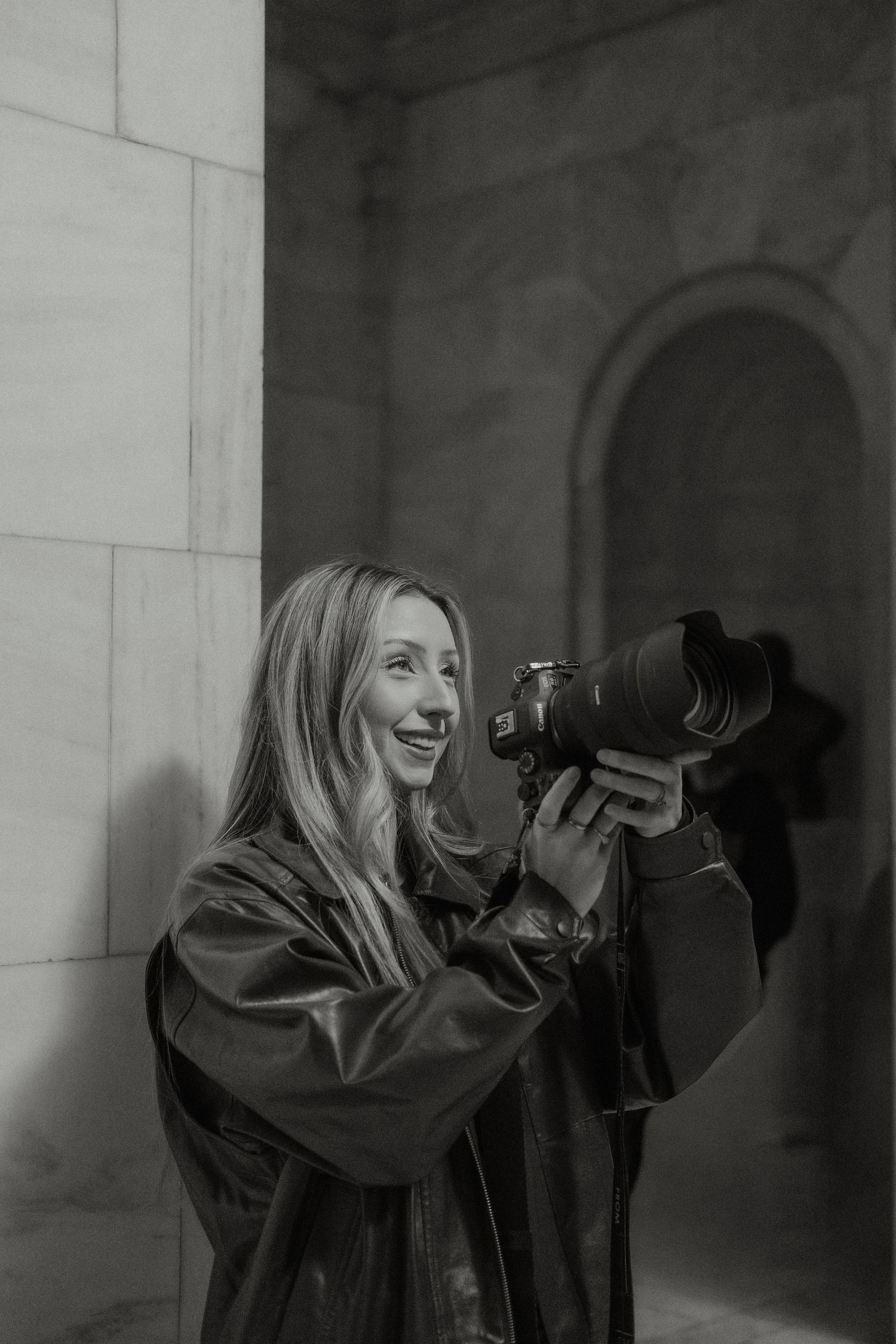 Smiling woman photographer holding DSLR camera against stone wall in black and white portrait.
