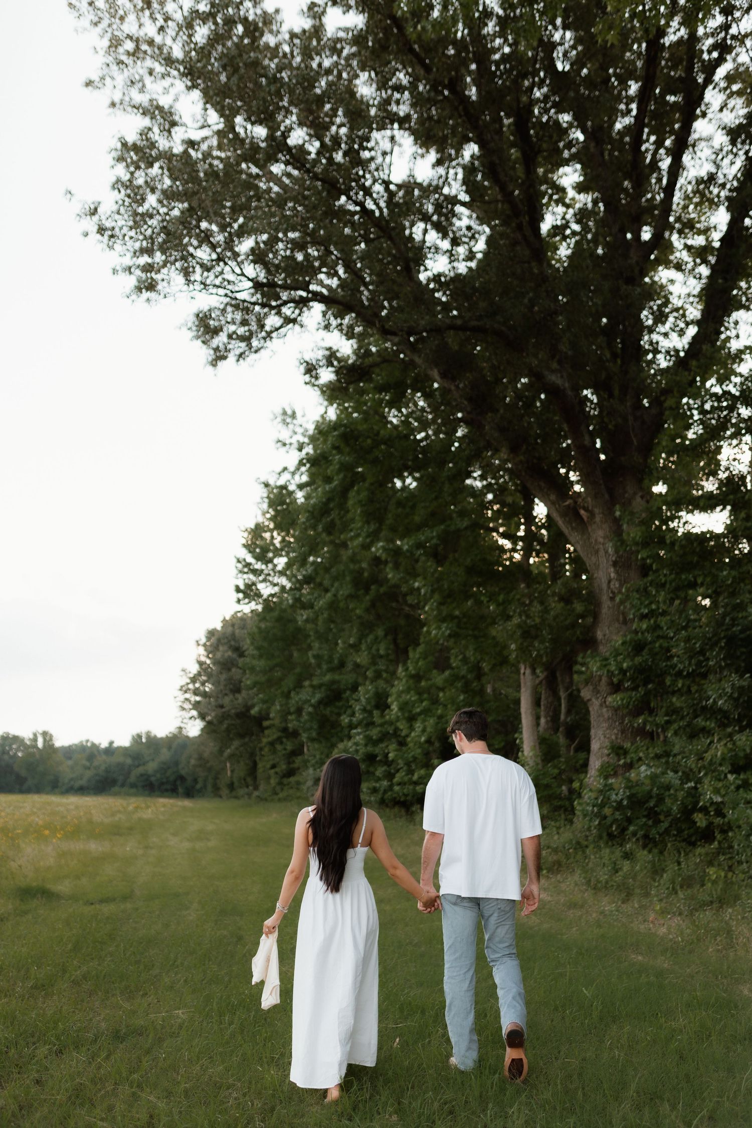 Couple holding hands walking toward trees in a green meadow, woman in flowing white dress, man in white tee and jeans.