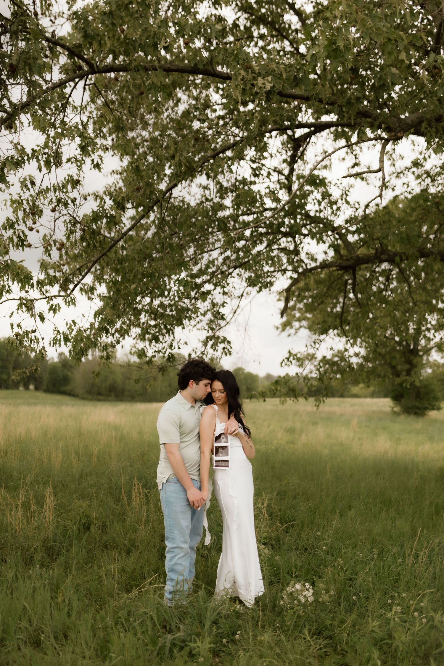 Couple standing in green field under large tree, woman in white dress holding ultrasound photos during maternity shoot.