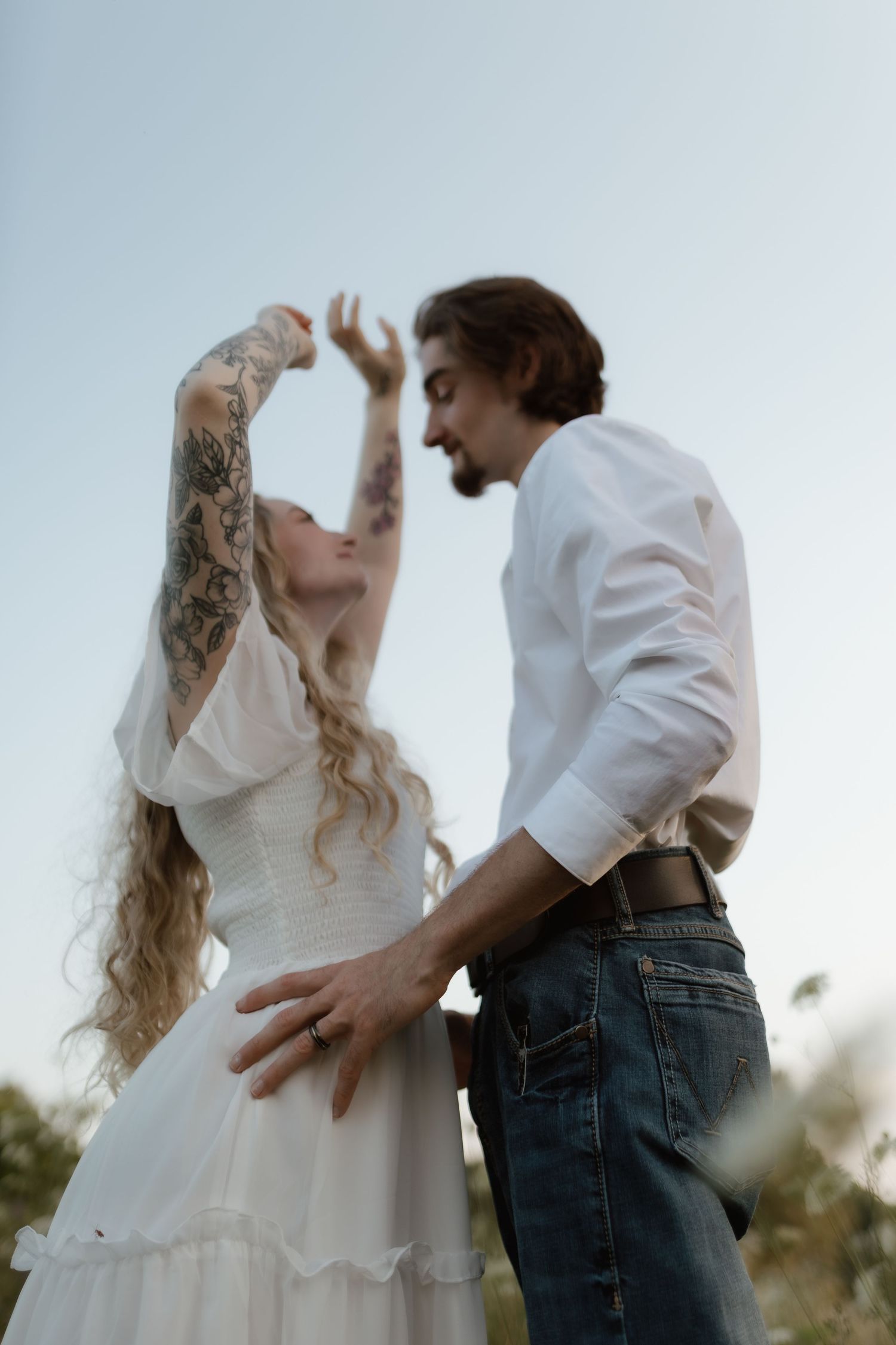 Tattooed woman in white dress reaching up while facing bearded man in white shirt in a field at dusk, close embrace.