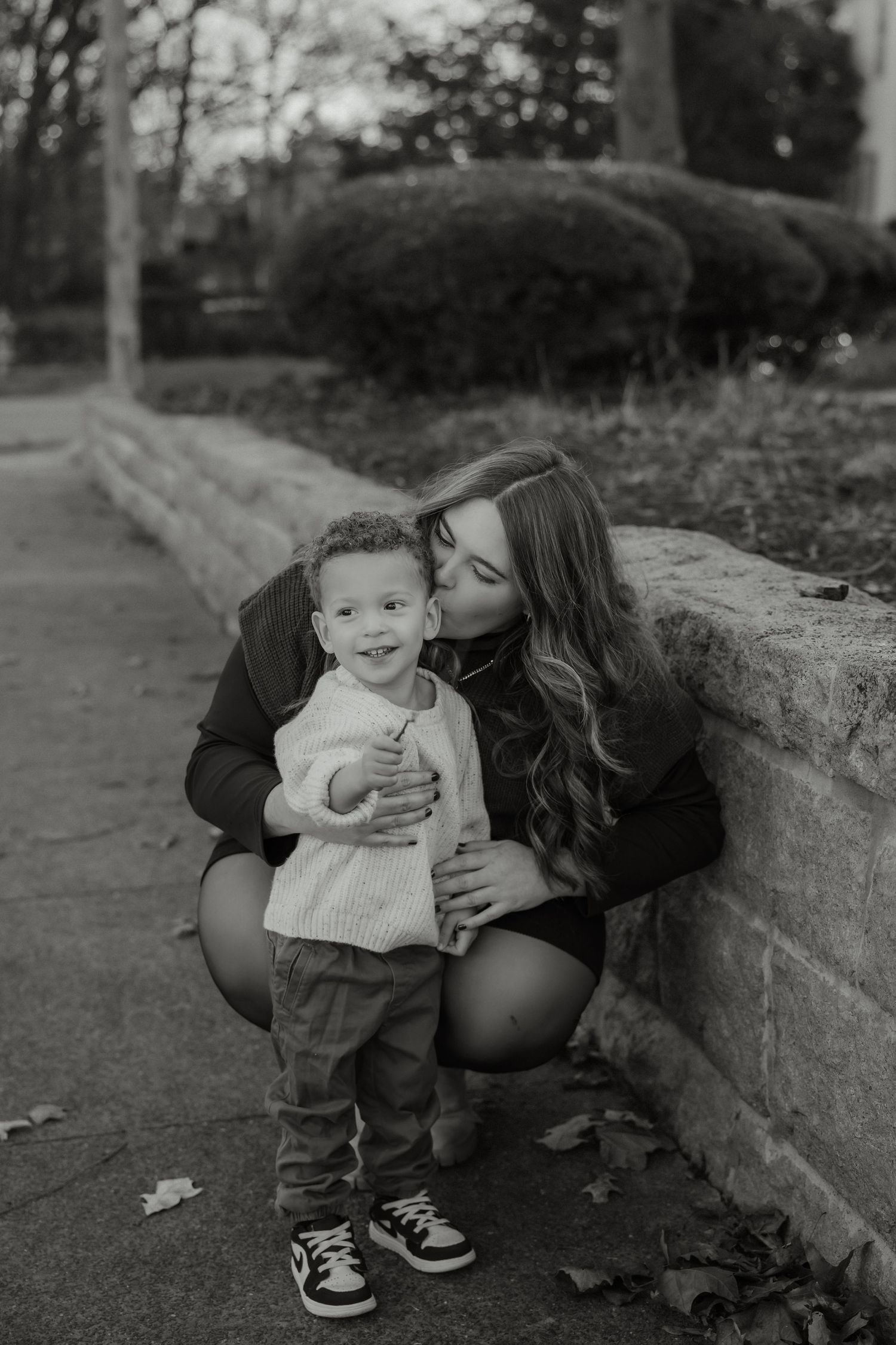 Black and white photo of mother kissing curly-haired toddler son leaning against stone wall.