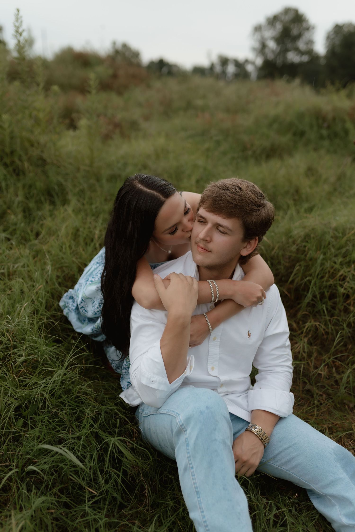 A couple sits in tall grass as the woman embraces the man from behind and kisses his cheek.