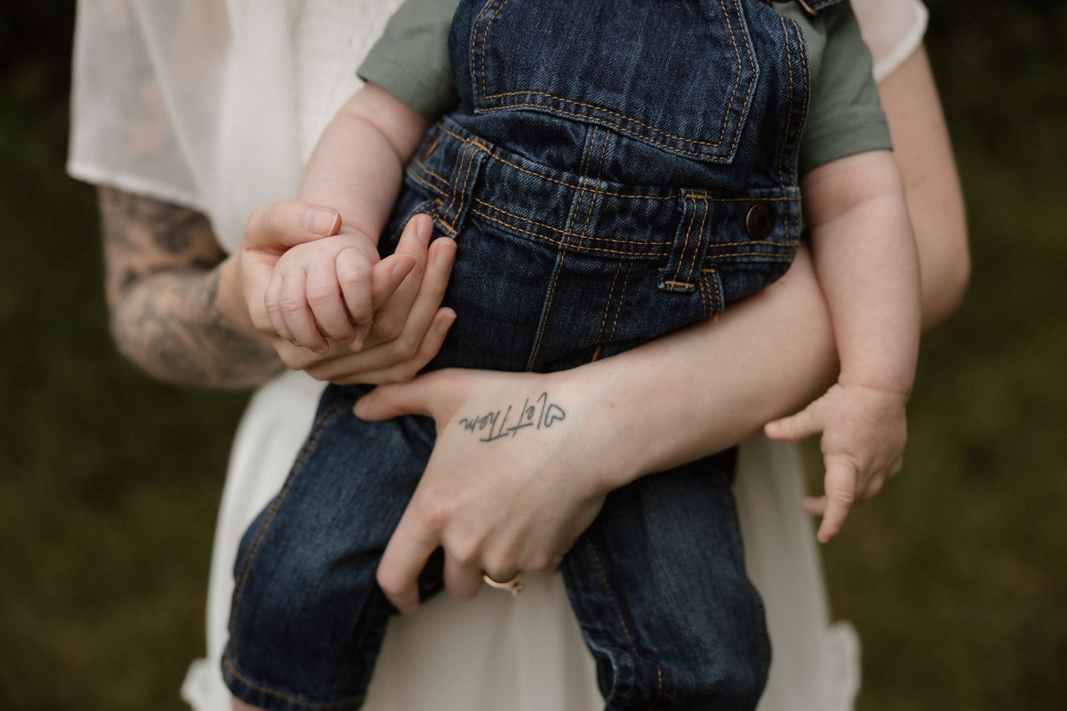 Mother with tattoo holding baby in denim overalls outdoors.