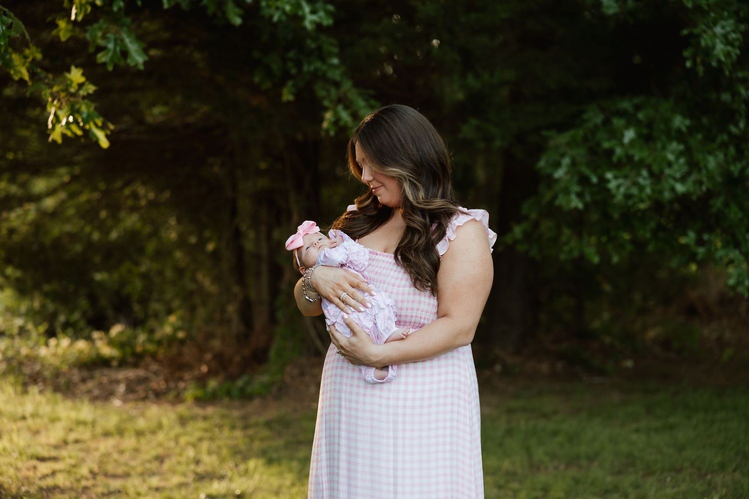 Brunette mother in gingham dress cradling newborn in green park.