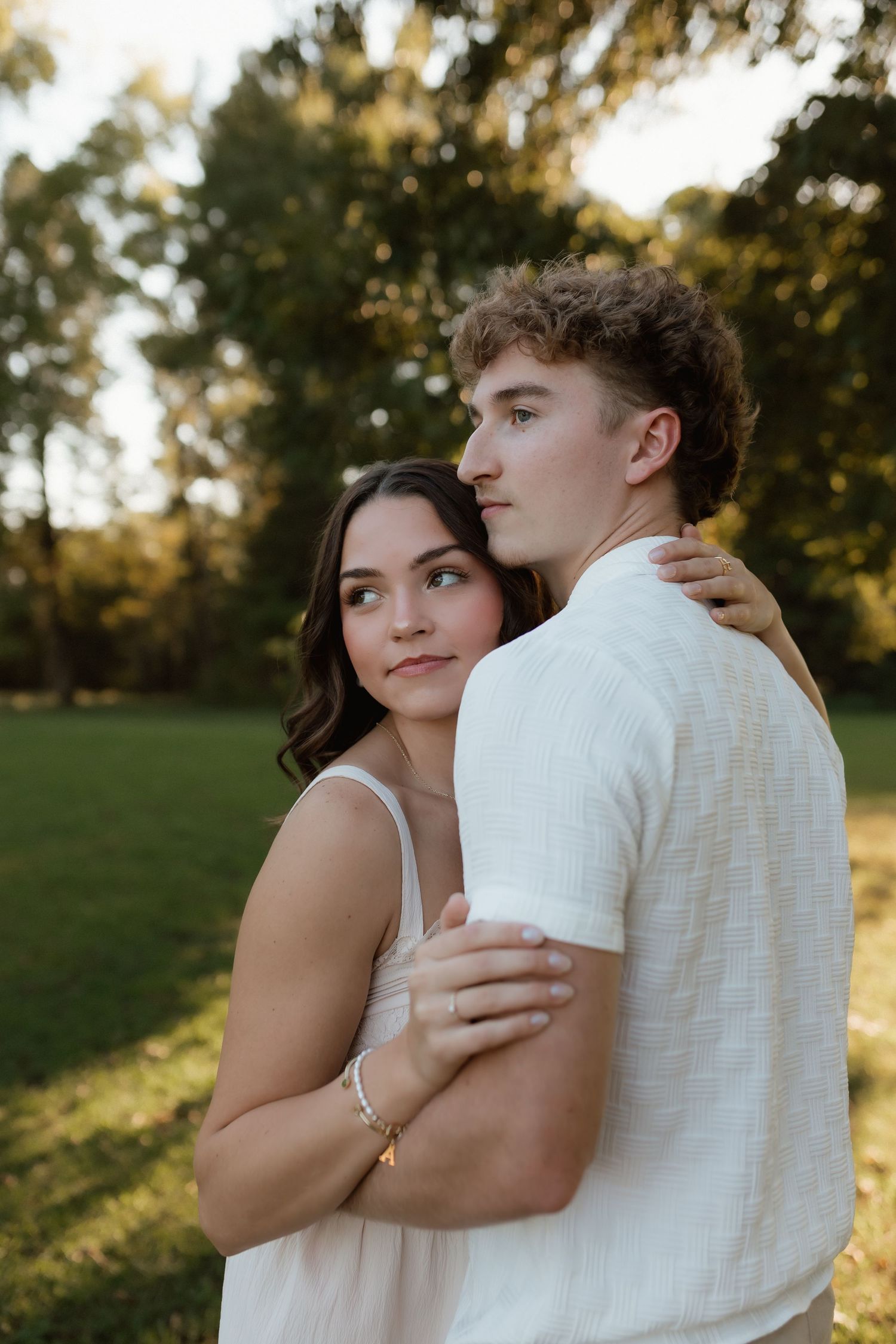 A young couple embraces in a sunlit park at golden hour, woman in white dress hugging man from behind peacefully.