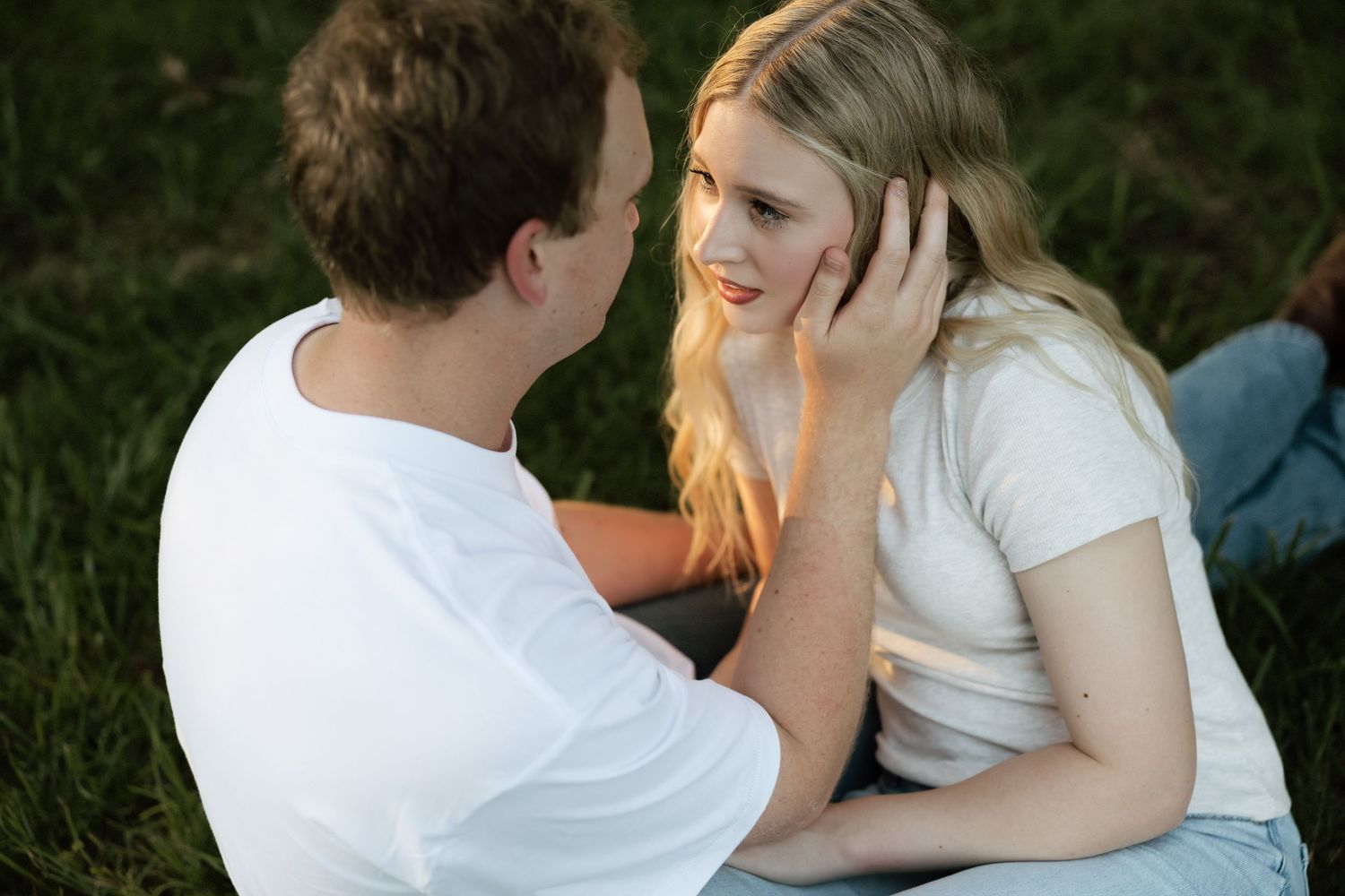 Tender moment as man gently touches blonde womans face while sitting together in green grass at sunset.