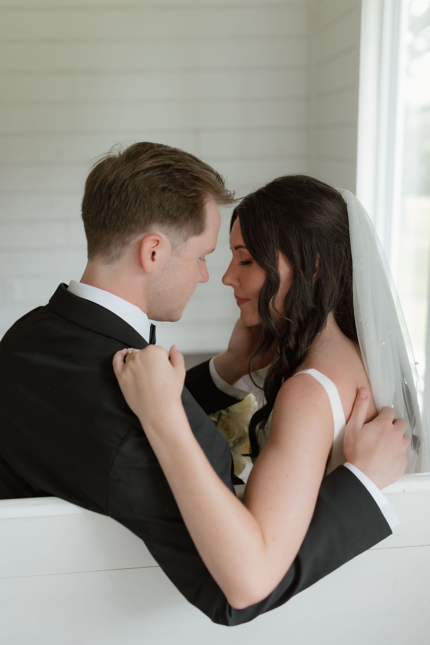 Bride and groom sharing an intimate moment together near a bright window inside a white chapel.