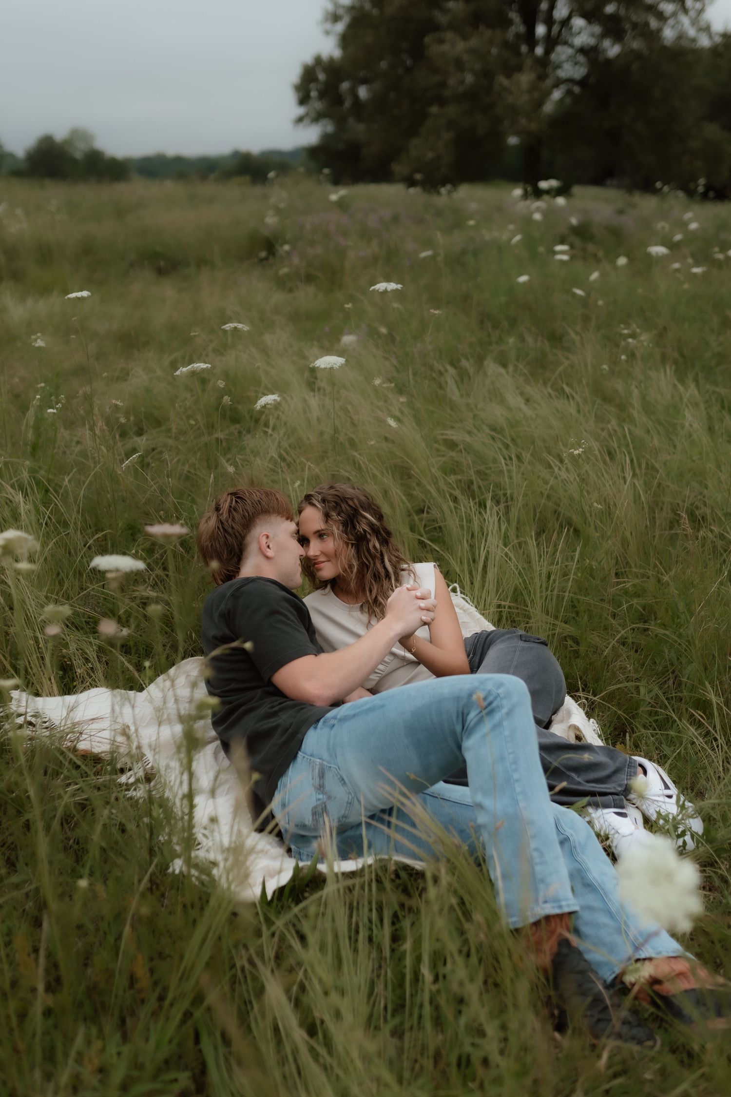 Couple sitting together on blanket in wildflower meadow, holding mugs and leaning in for a kiss.