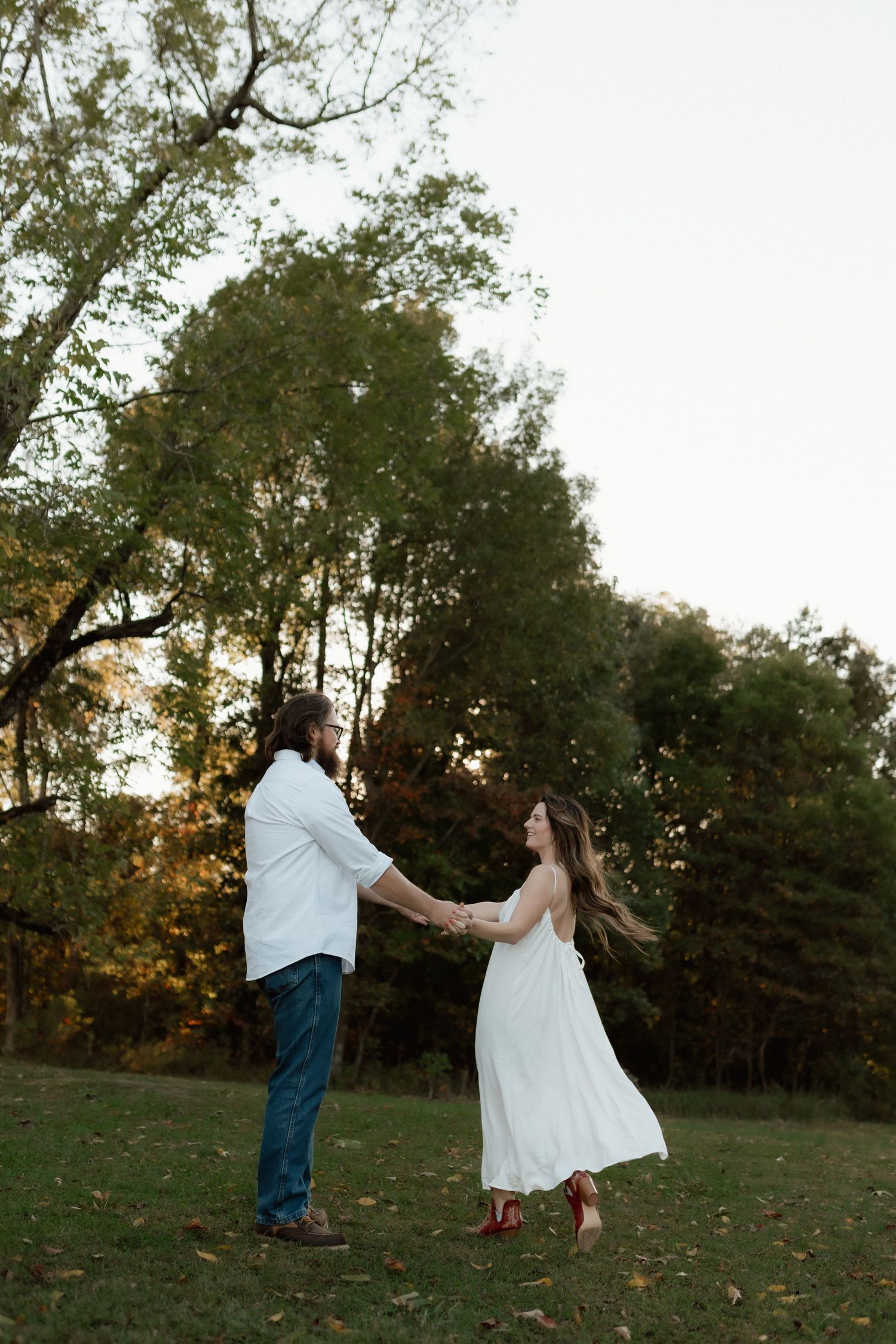 A couple holds hands and dances playfully in a lush green park at sunset, woman wearing white dress and red boots.
